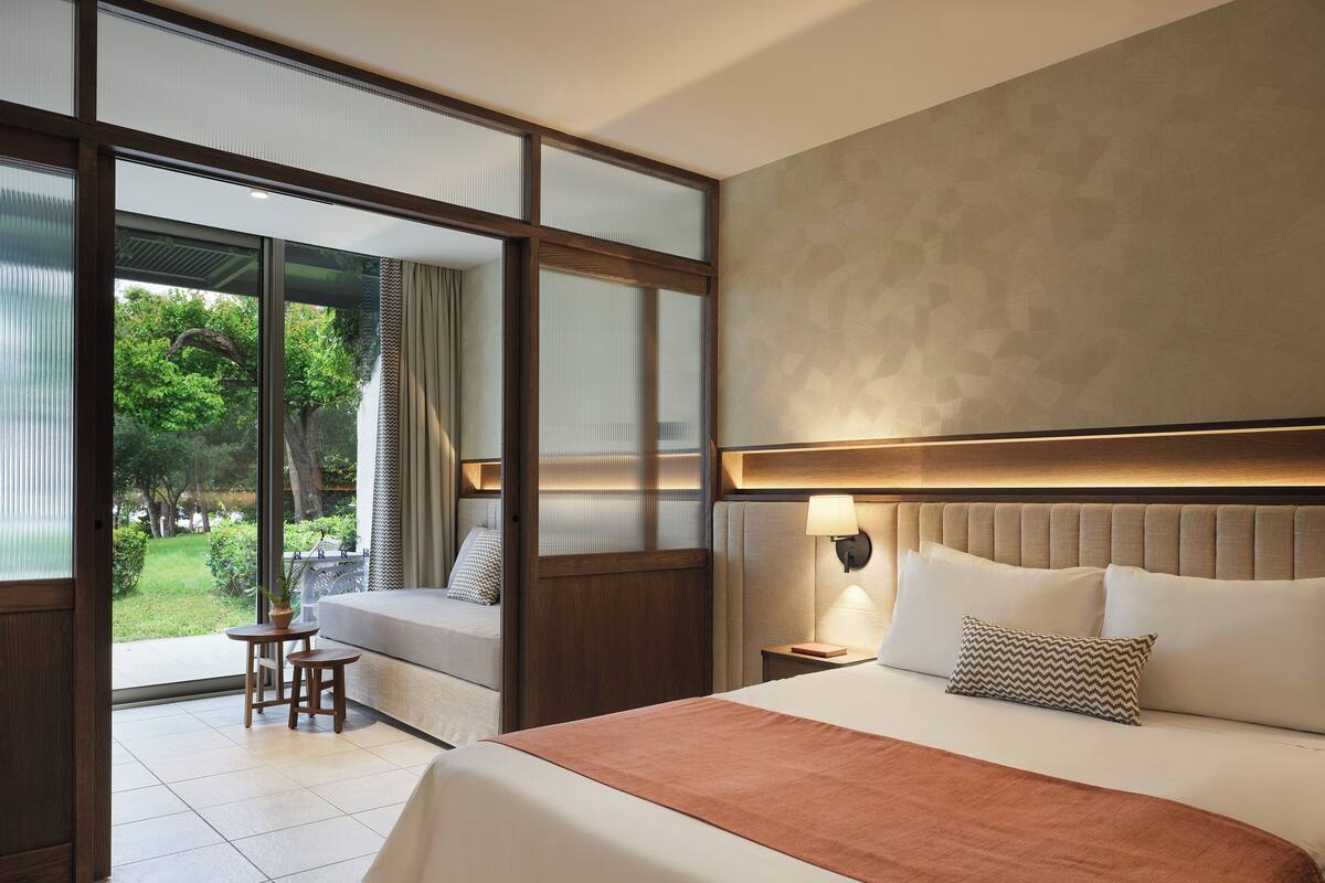 A hotel room with a large bed, a nightstand with a lamp, a view of a garden through a sliding door, and a small seating area with a table and chair.