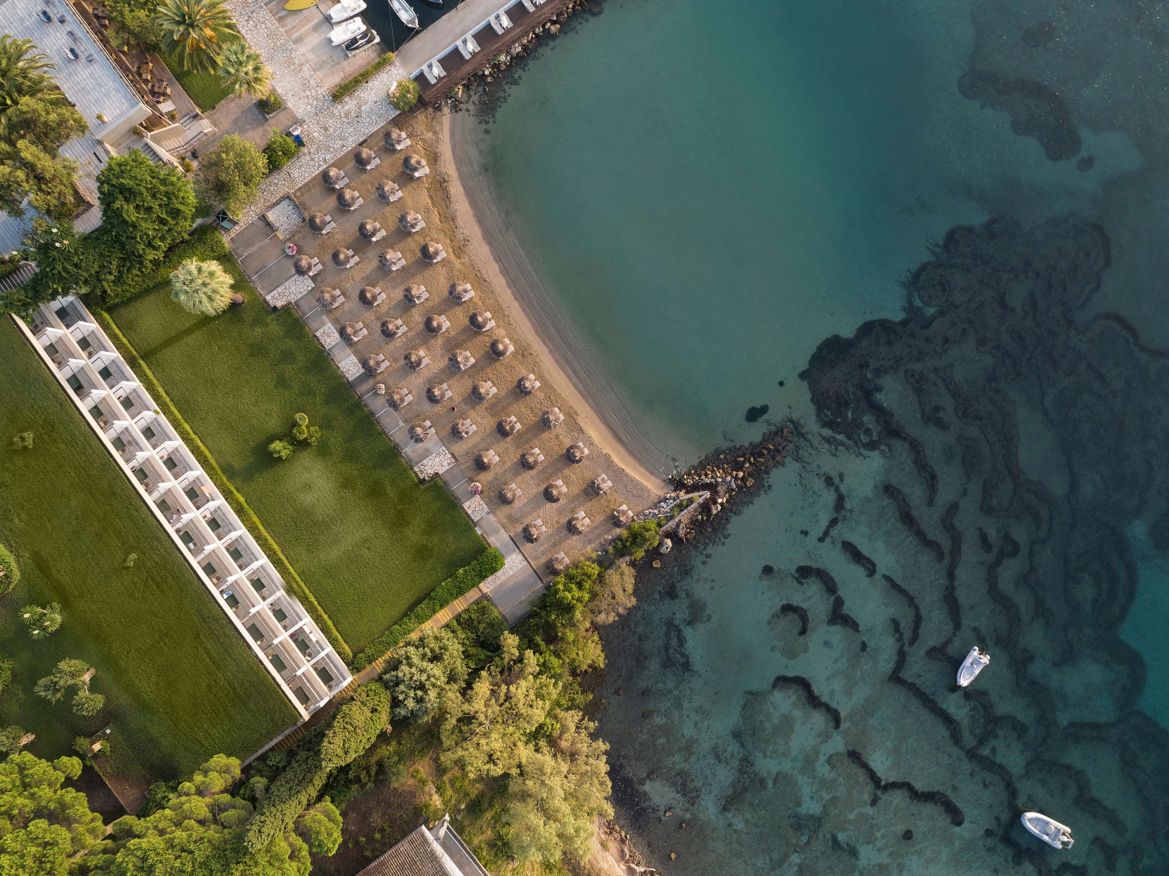 An aerial view of a coastal hotel with a long row of rooms, a beach area with umbrellas, and clear blue waters.