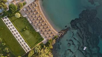Vue aérienne d’un hôtel situé sur la côte, avec une longue rangée de chambres, une plage agrémentée de parasols et une eau d’un bleu limpide.