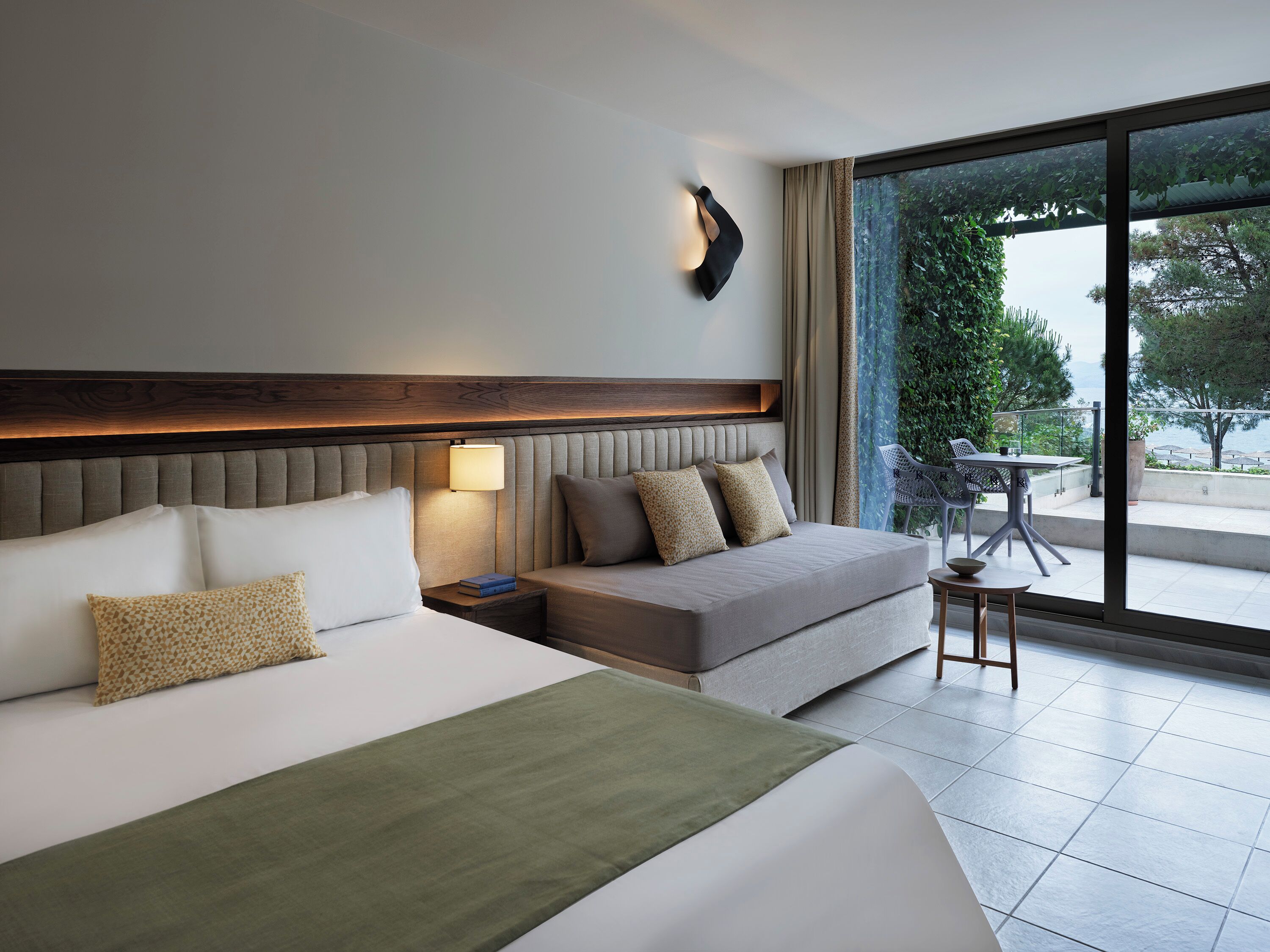 A hotel room with a large bed, a bedside table, a lamp, and a view of the outdoors through a large window.