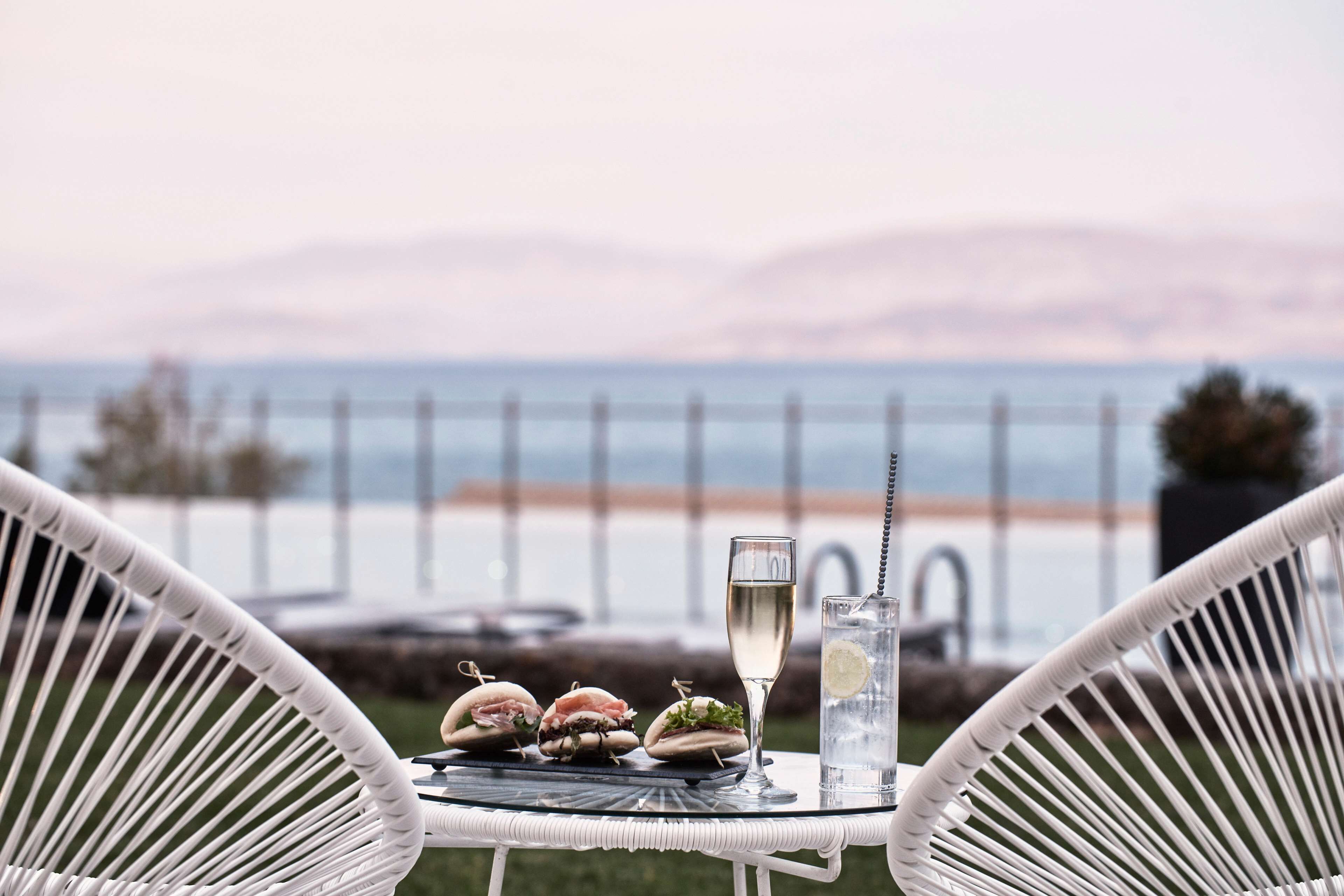 Uma mesa com três sanduíches e uma taça de champanhe, com vista para o mar.