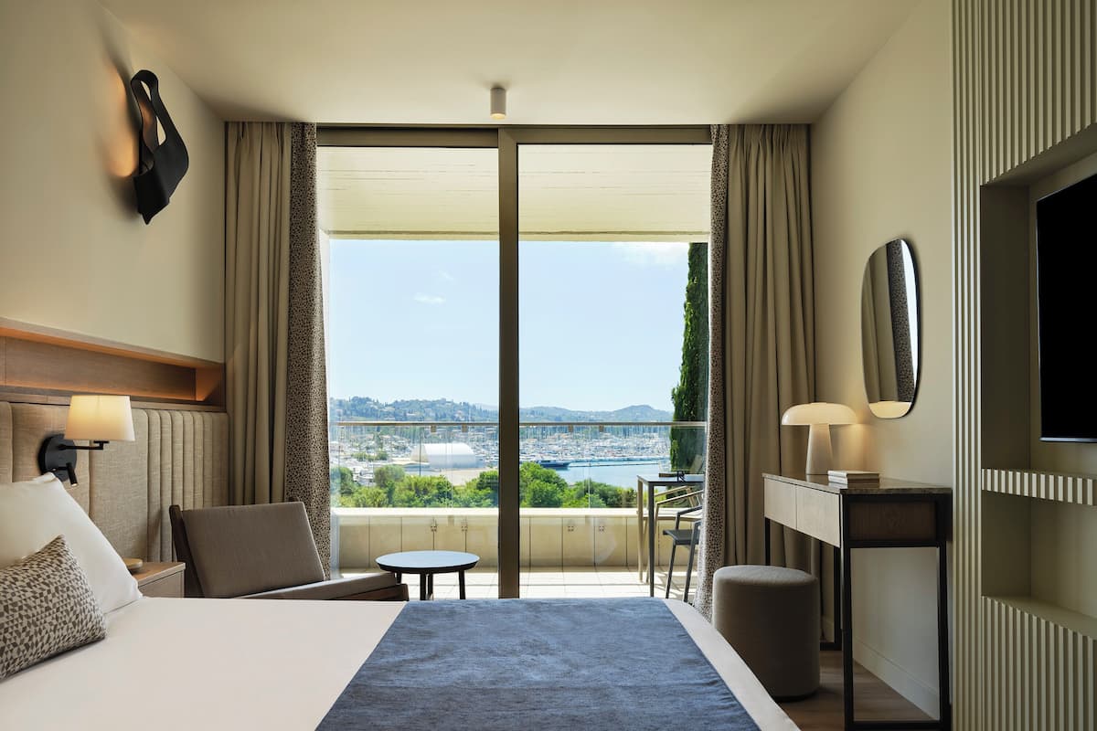 A hotel room with a large bed, a desk, a chair, and a view of a cityscape through a large window.