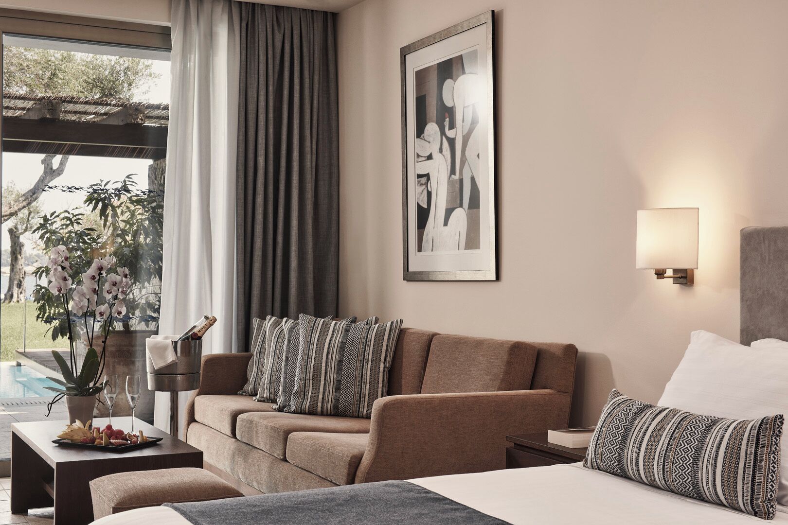 A hotel room with a sofa, a bed, a nightstand, a lamp, a framed artwork, and a view of the outdoors.