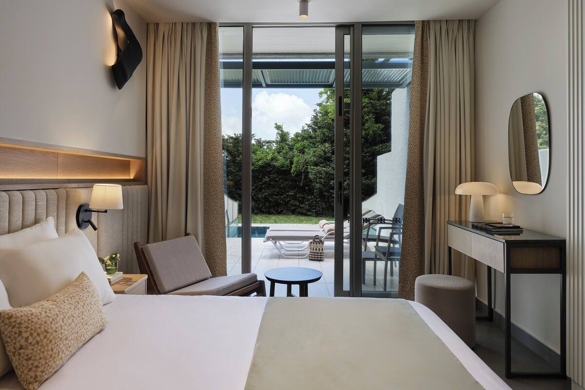 A hotel room with a bed, a desk, a chair, a small table, and a view of a pool and garden.