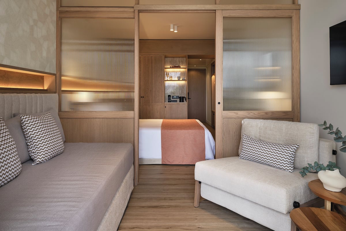 A modern hotel room with a sofa, a chair, a bed, and a kitchenette area.