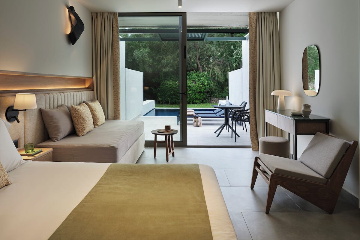 A hotel room with a bed, a desk, a chair, a small table, and a view of a pool and garden.