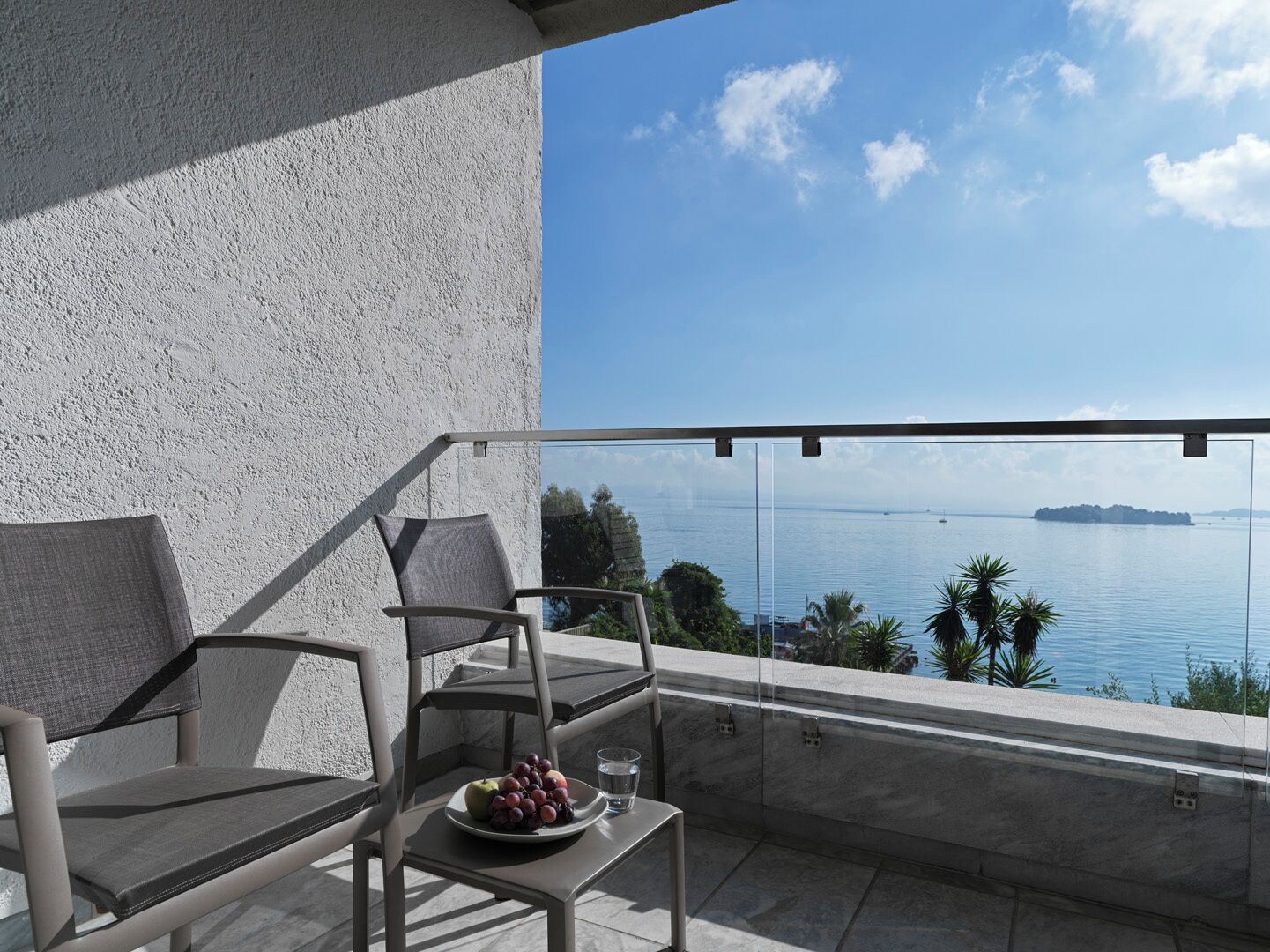 Varanda com duas cadeiras, uma mesa pequena com uma travessa de fruta, e vista desobstruída para o mar e ilhas ao longe.