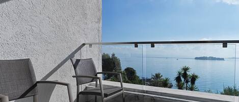 A balcony with two chairs, a small table with a plate of fruit, and a clear view of the sea and distant islands.