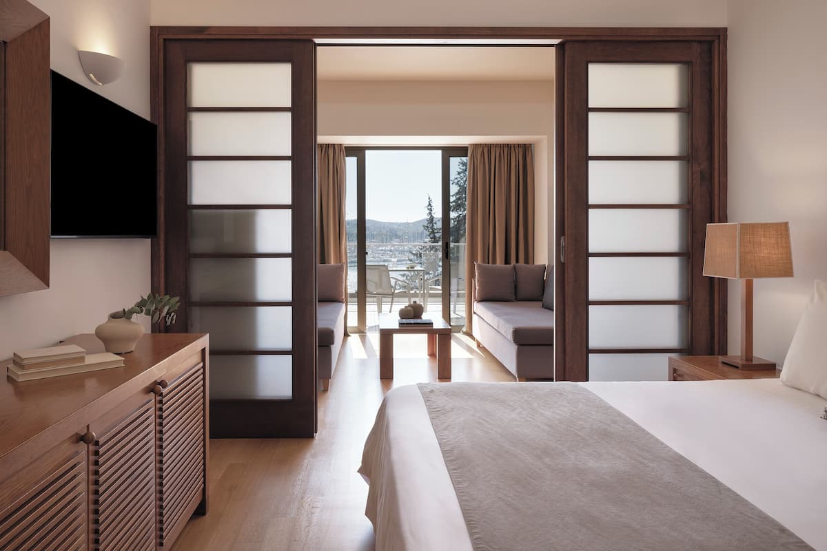 A hotel room with a large bed, a desk with a lamp, a sofa, and a view of the city through a glass door.