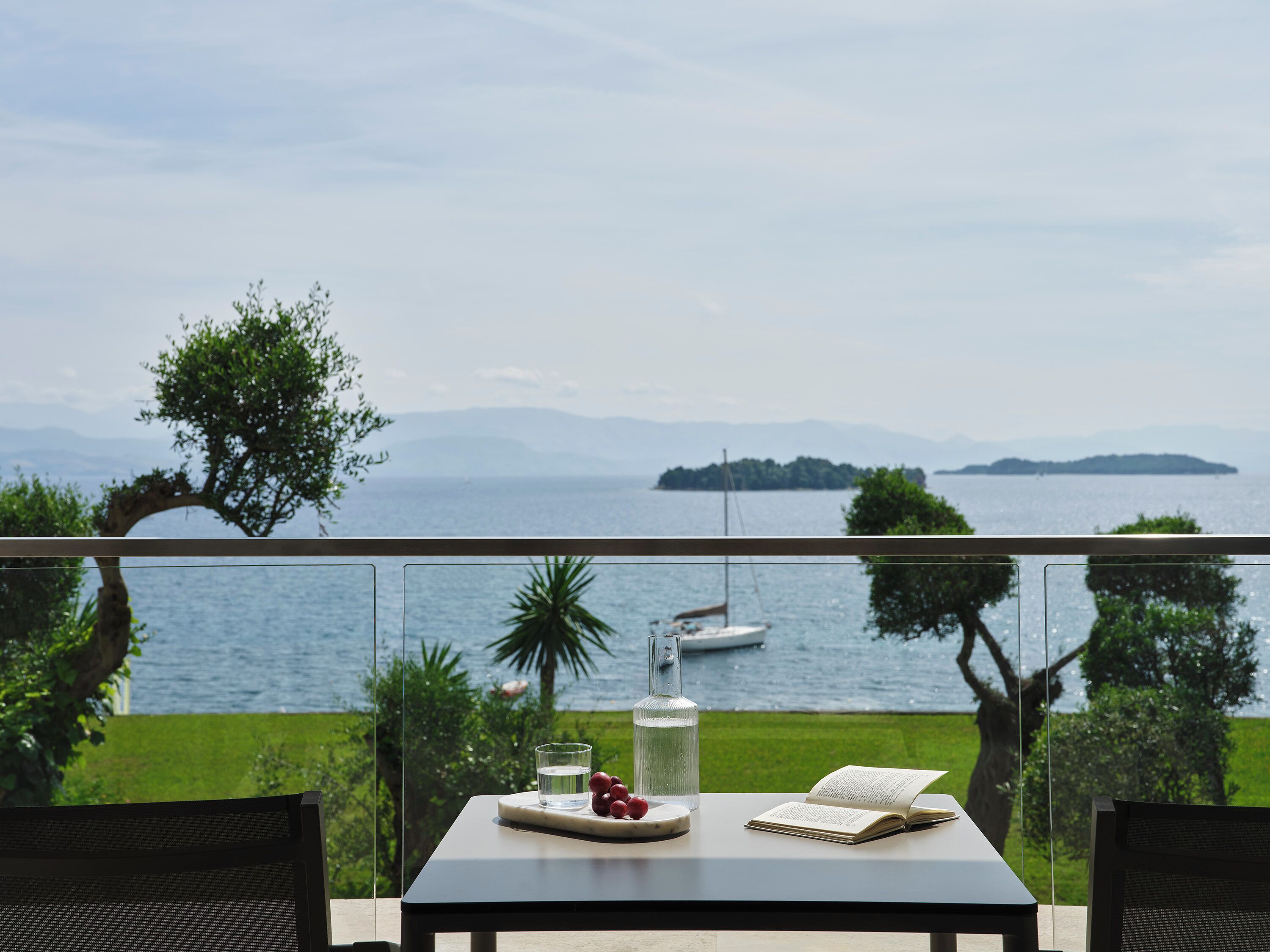 A table with a book, a glass of water, and a bottle on a balcony overlooking a body of water with boats and islands.