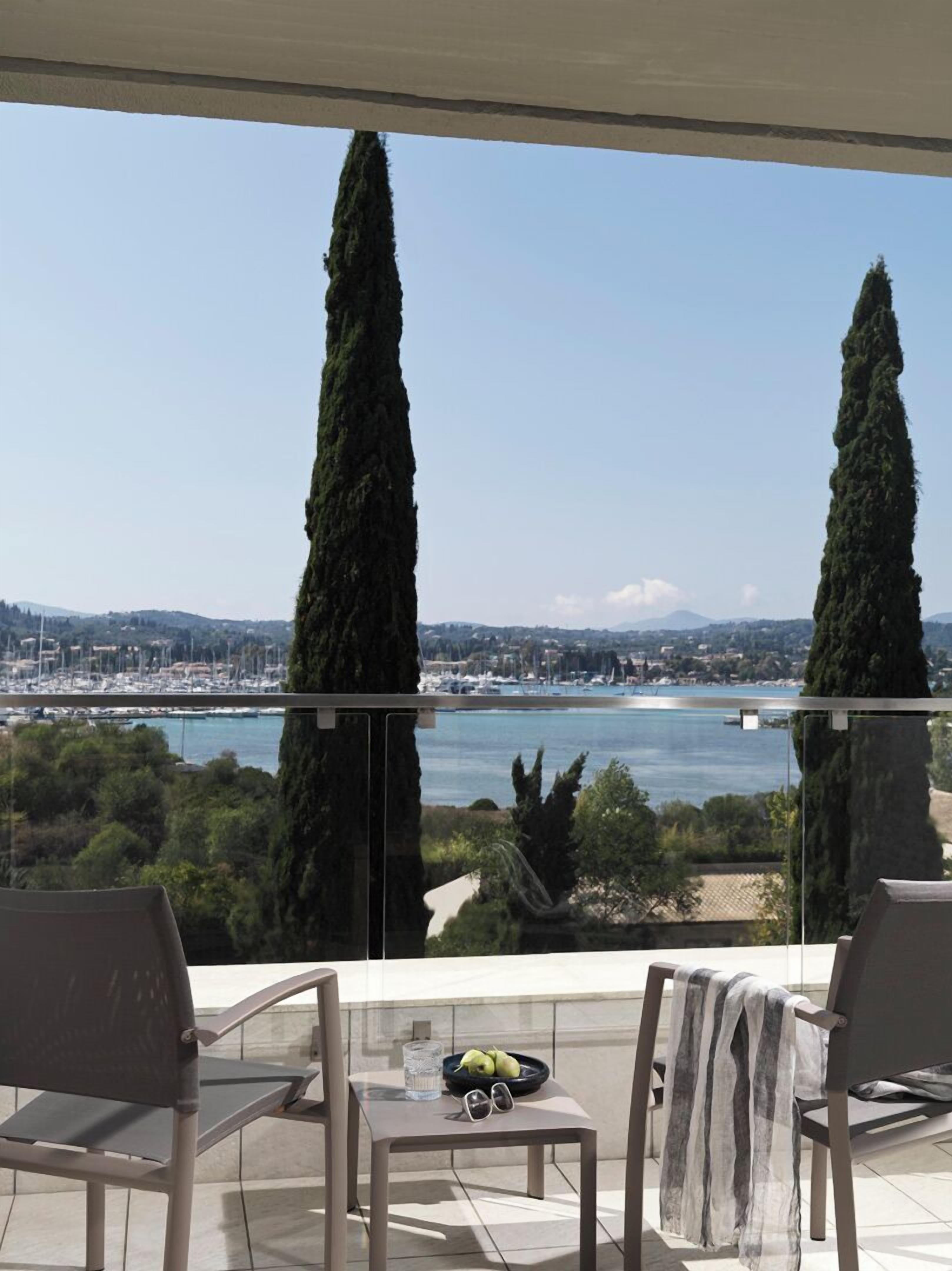 A balcony with two chairs, a table with a bowl of fruit and a glass of water, and a view of a marina and cityscape.
