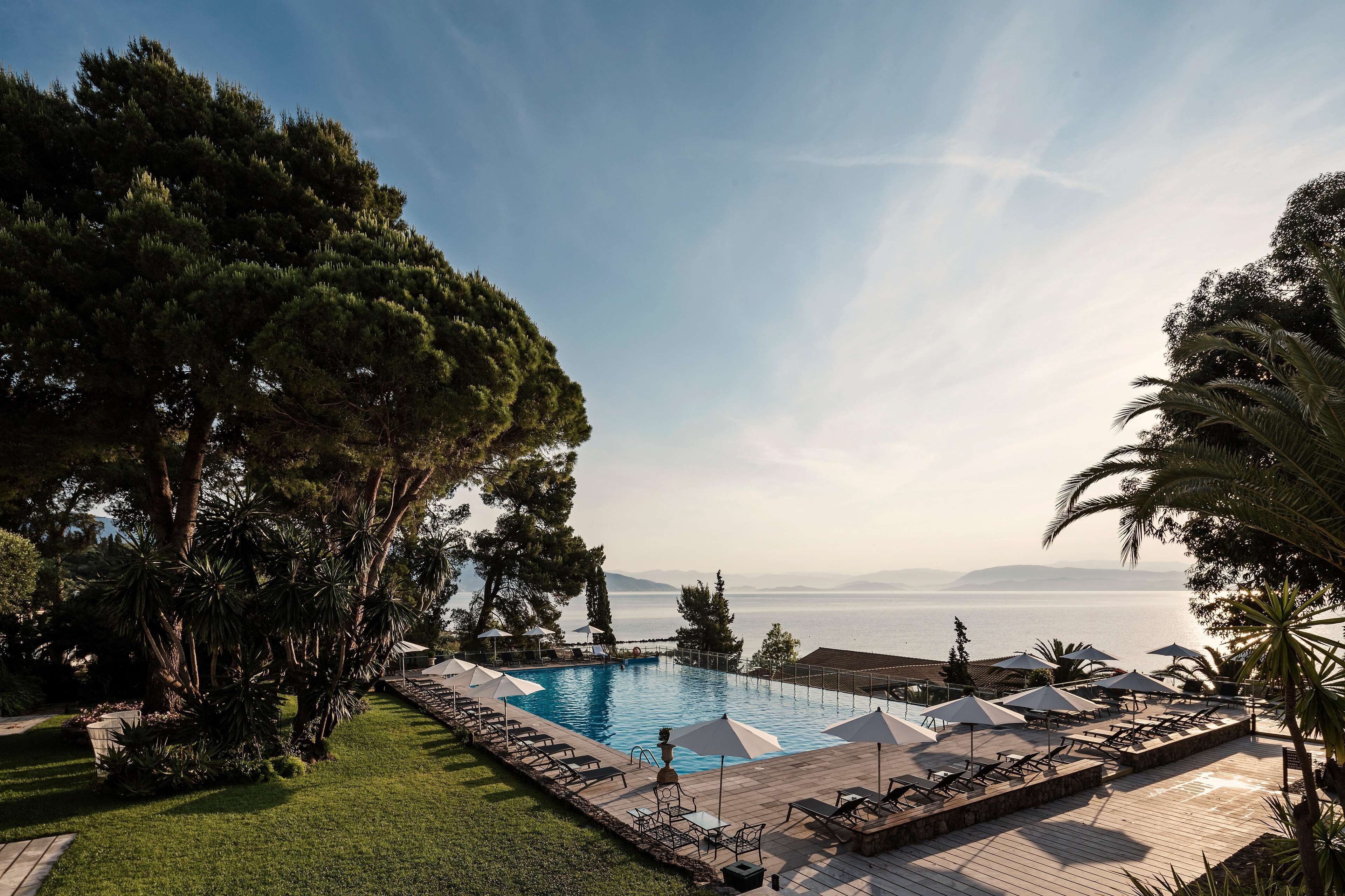 Ein Poolbereich mit Liegestühlen und Sonnenschirmen, umgeben von Bäumen und mit Blick auf ein Gewässer.