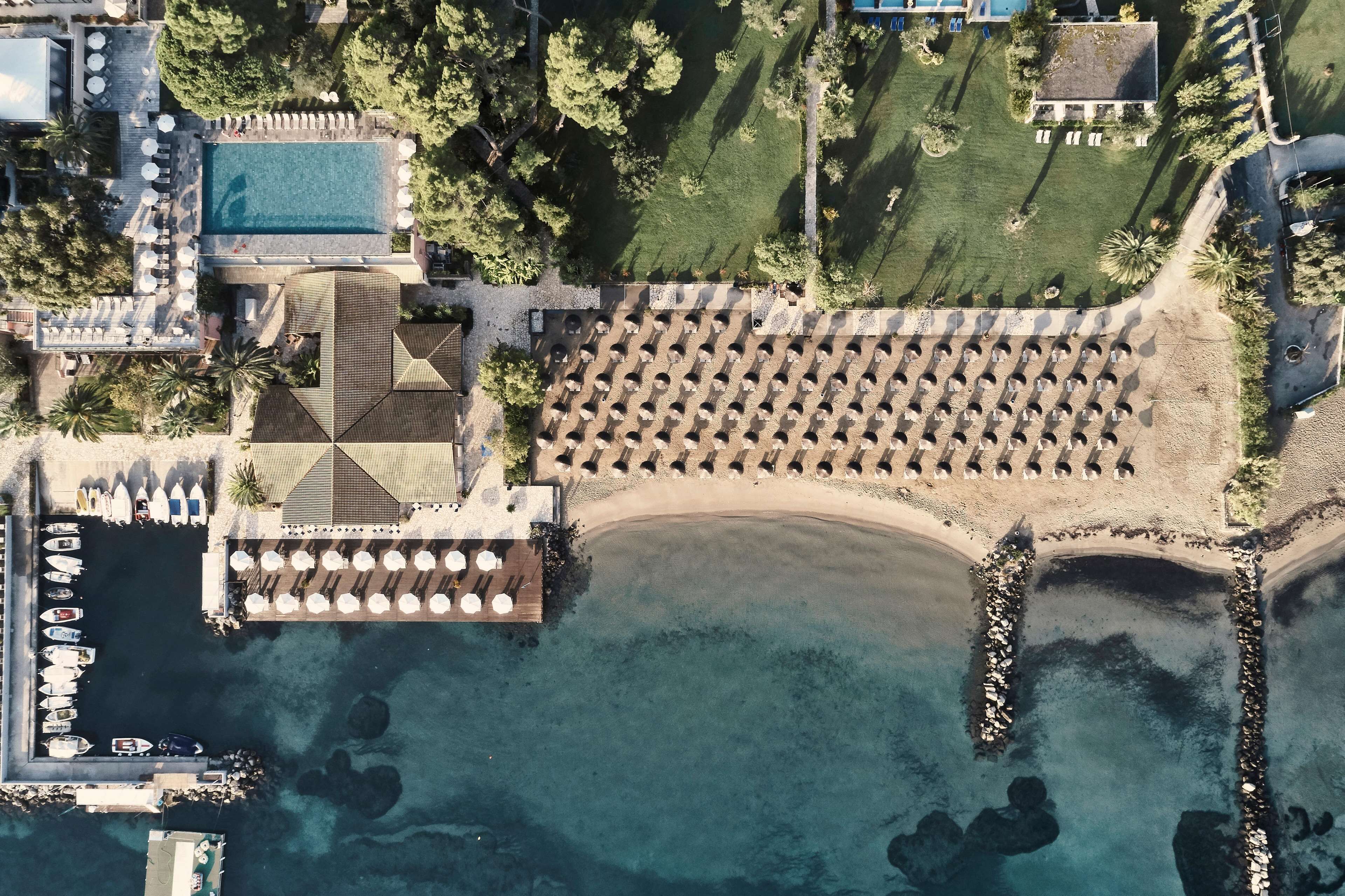 Luftaufnahme eines Resorts mit Schwimmbad, Strandbereich und Reihen von Sonnenschirmen.