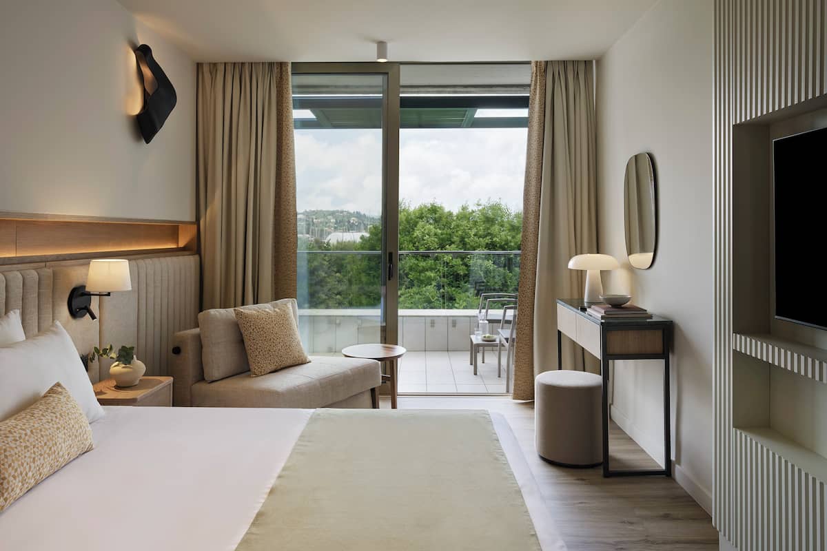 A hotel room with a large bed, a sofa, a desk with a lamp, a TV, and a balcony with a view of greenery.