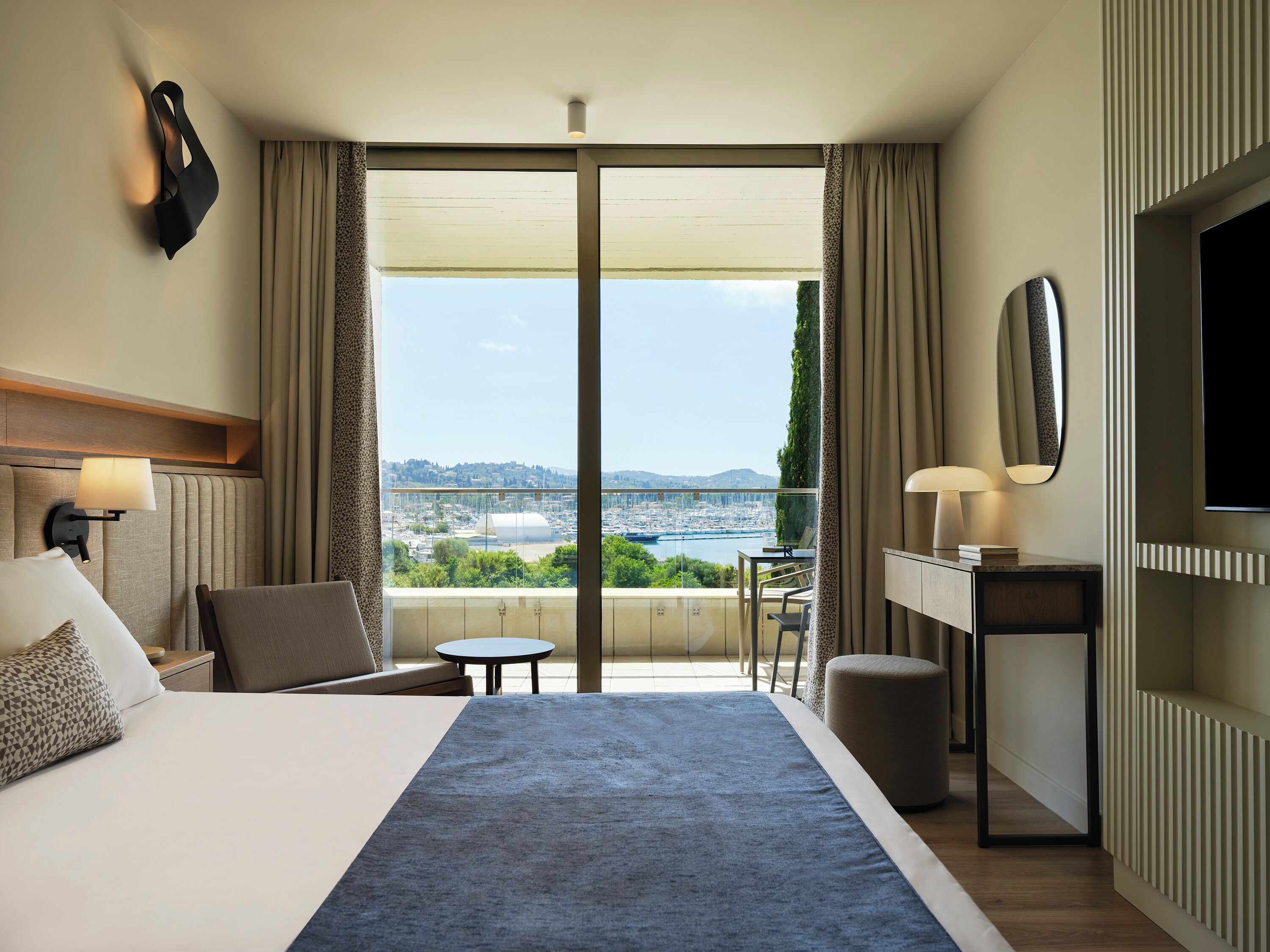 A hotel room with a large bed, a desk, a chair, and a view of a cityscape through a large window.