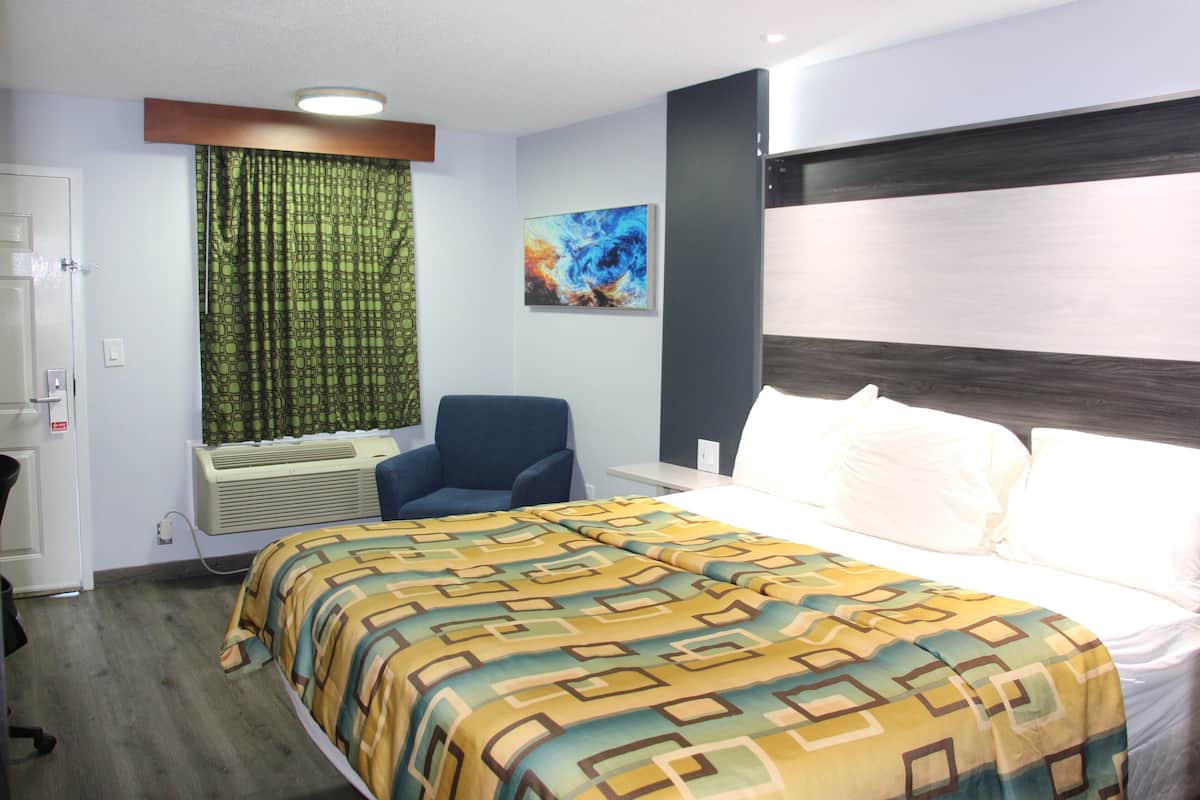 A hotel room with a bed, a chair, a window with curtains, a painting on the wall, and a wall-mounted air conditioner.