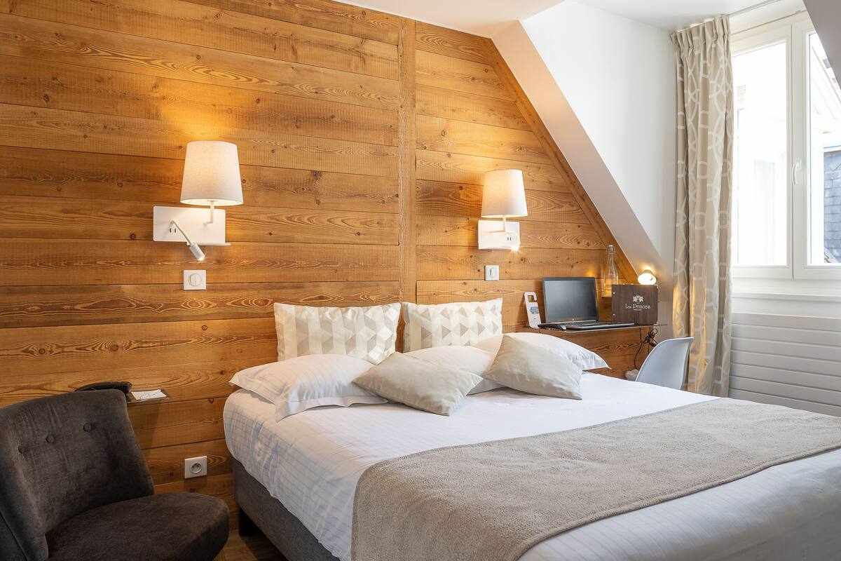 A bedroom with a wooden wall, a bed with white bedding, two bedside lamps, a chair, and a desk with a lamp.