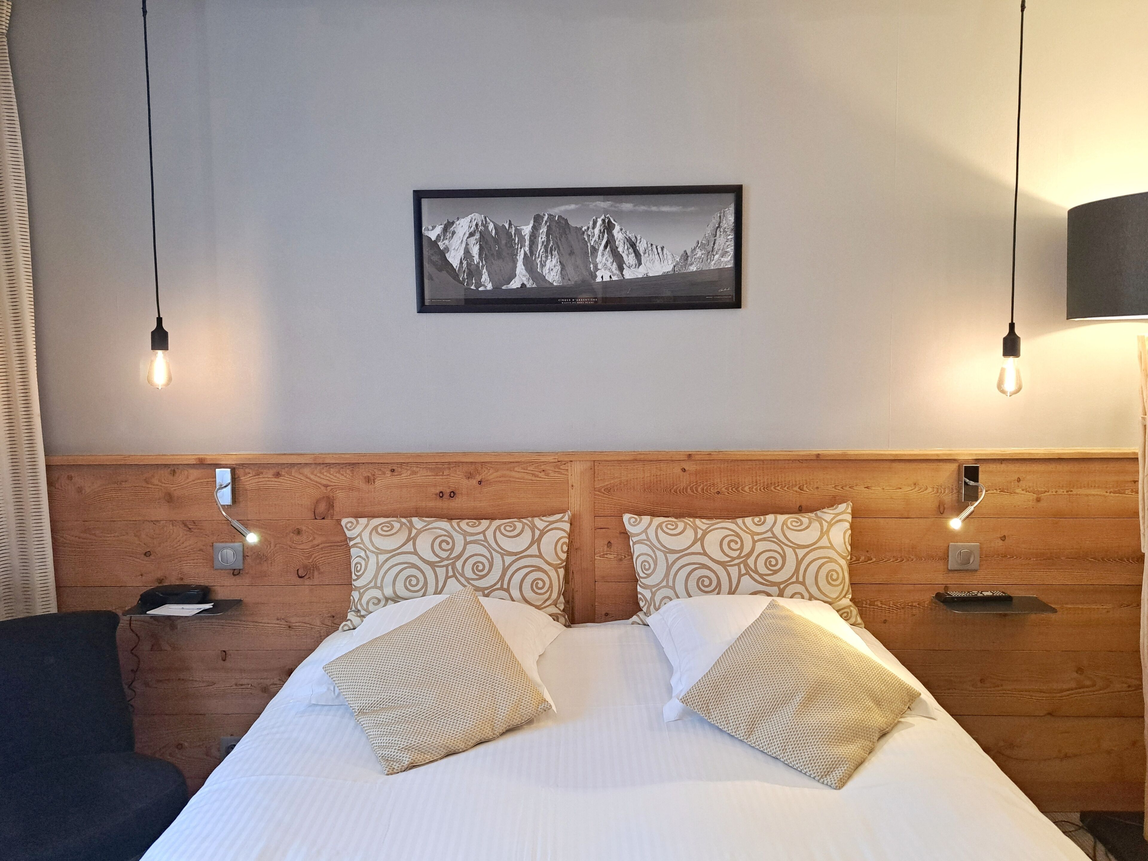 A hotel room with a wooden headboard, two patterned pillows, a framed mountain landscape picture, and bedside lamps.