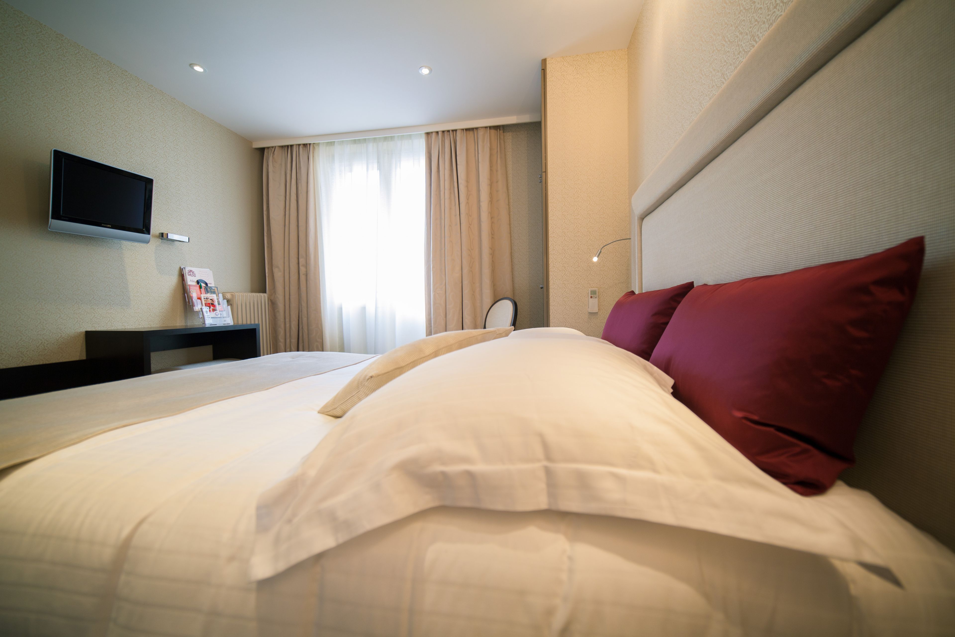 A hotel room with a bed, a flat-screen TV mounted on the wall, a bedside table, and a window with curtains.