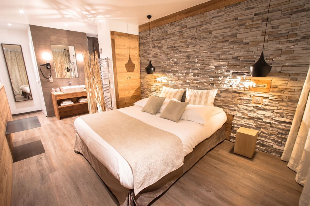 A bedroom with a stone wall, a wooden headboard, a bed with pillows, a nightstand, a mirror, and a wooden bench.