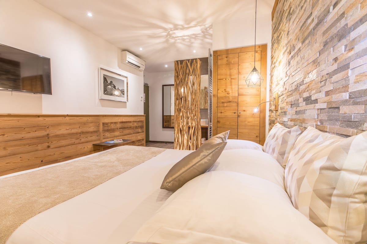 A modern bedroom with a large bed, wooden paneling, a mounted TV, and a stone wall.