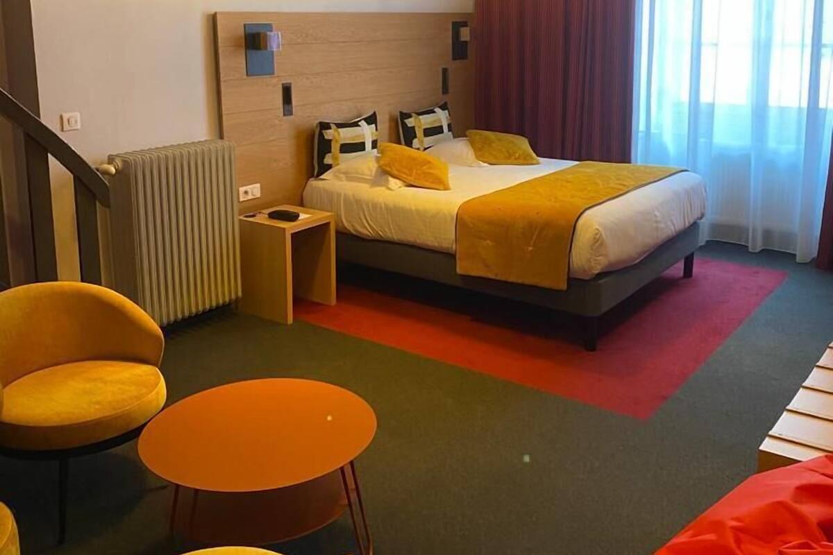 A hotel room with a bed, two bedside tables, a radiator, a chair, a small table, and a window with curtains.