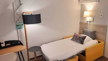 Une chambre d’hôtel de taille réduite, comprenant un lit, un bureau, une chaise, une lampe sur pied et un climatiseur fixé au mur.