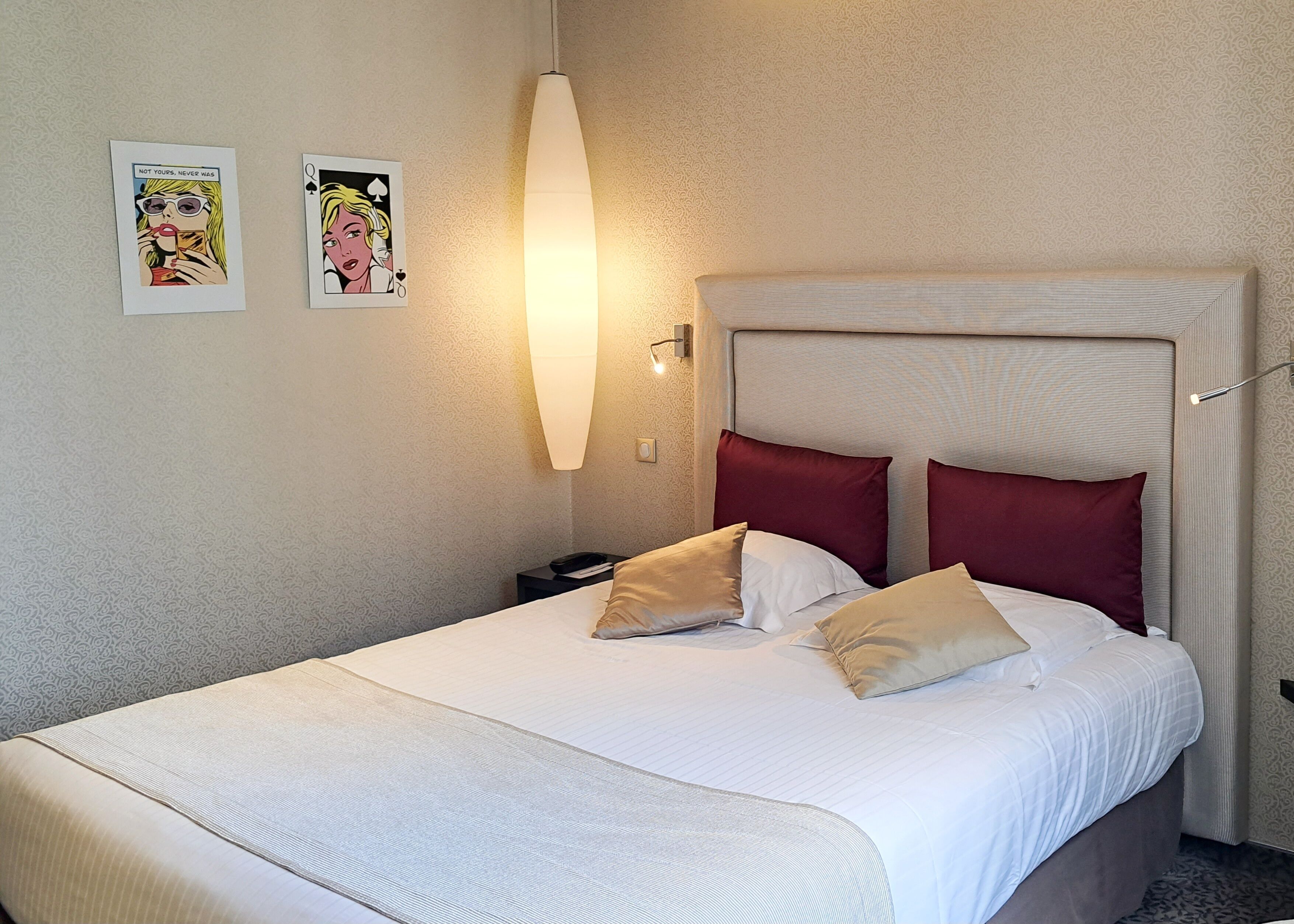 Une chambre d’hôtel avec un grand lit, deux oreillers rouges, une tête de lit blanche et deux œuvres d’art encadrées accrochées au mur.