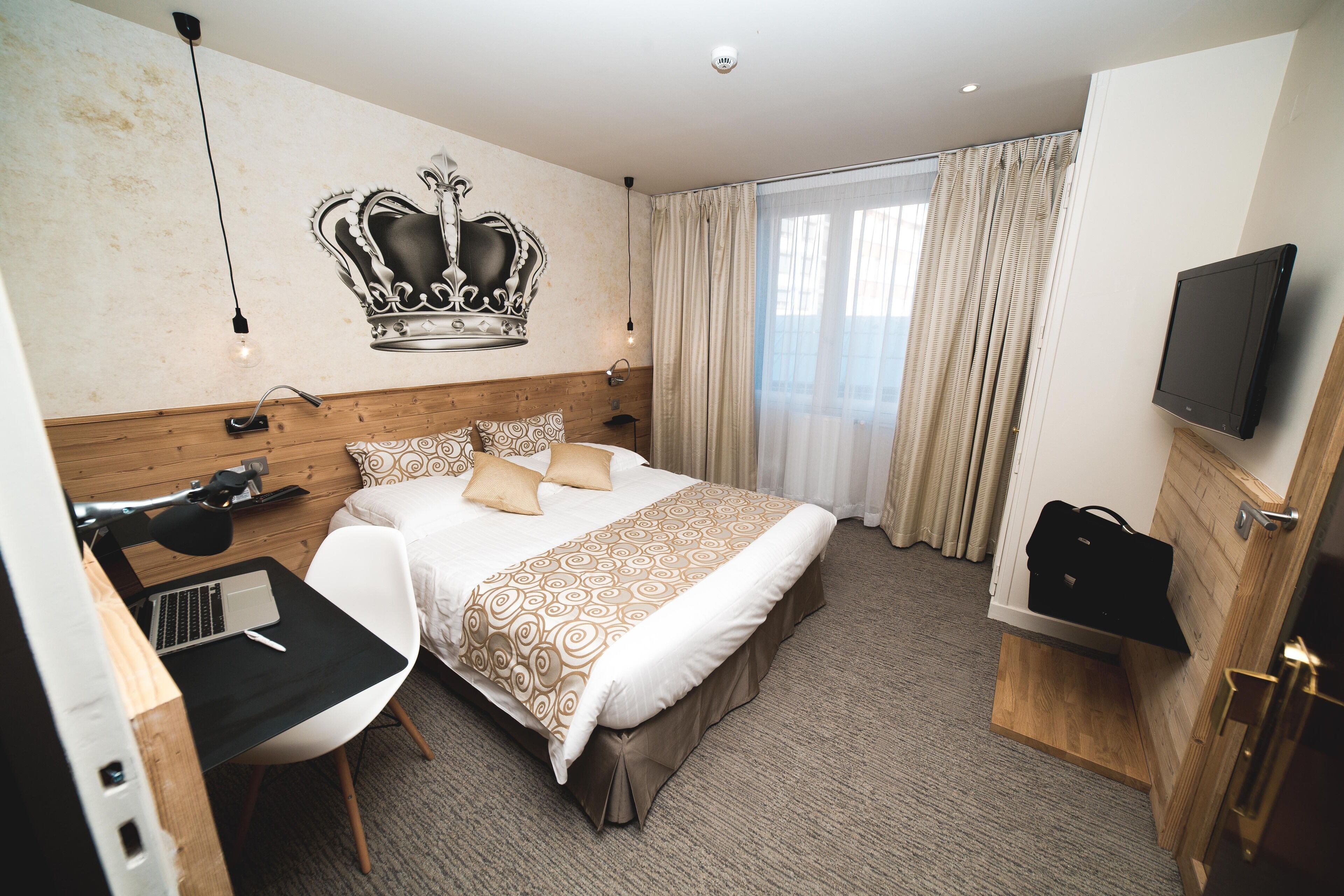 A hotel room with a bed, a desk with a laptop, a TV, and a crown mural on the wall.