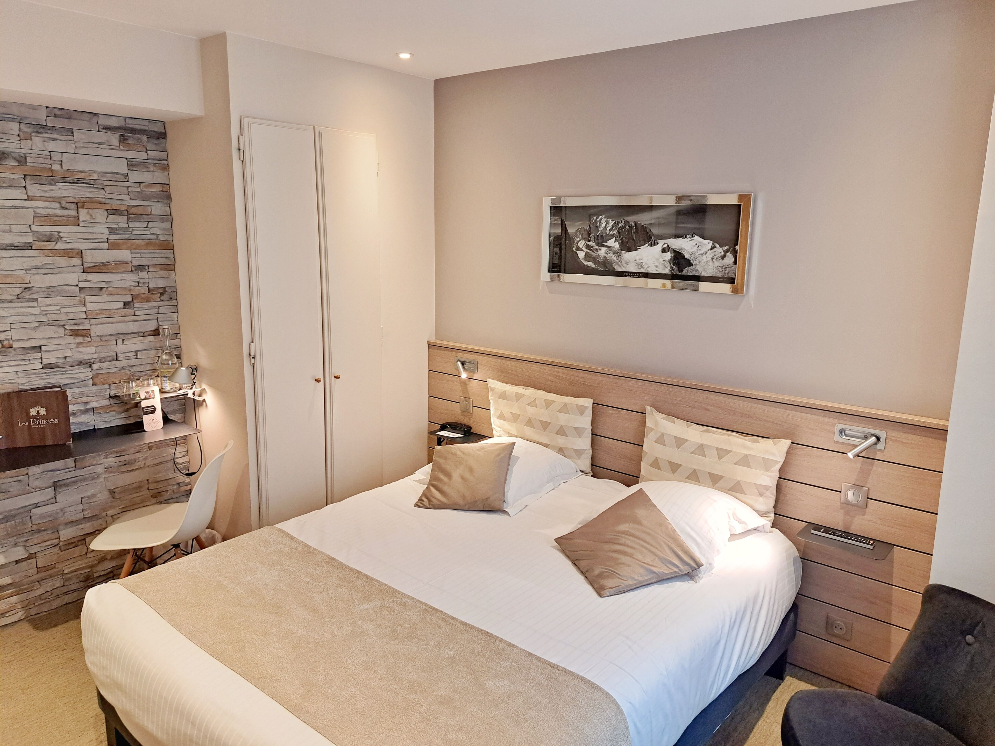 Un dormitorio con una cama grande, una mesita de noche, un cabecero de madera, una pared de piedra, una silla y un cuadro en la pared.