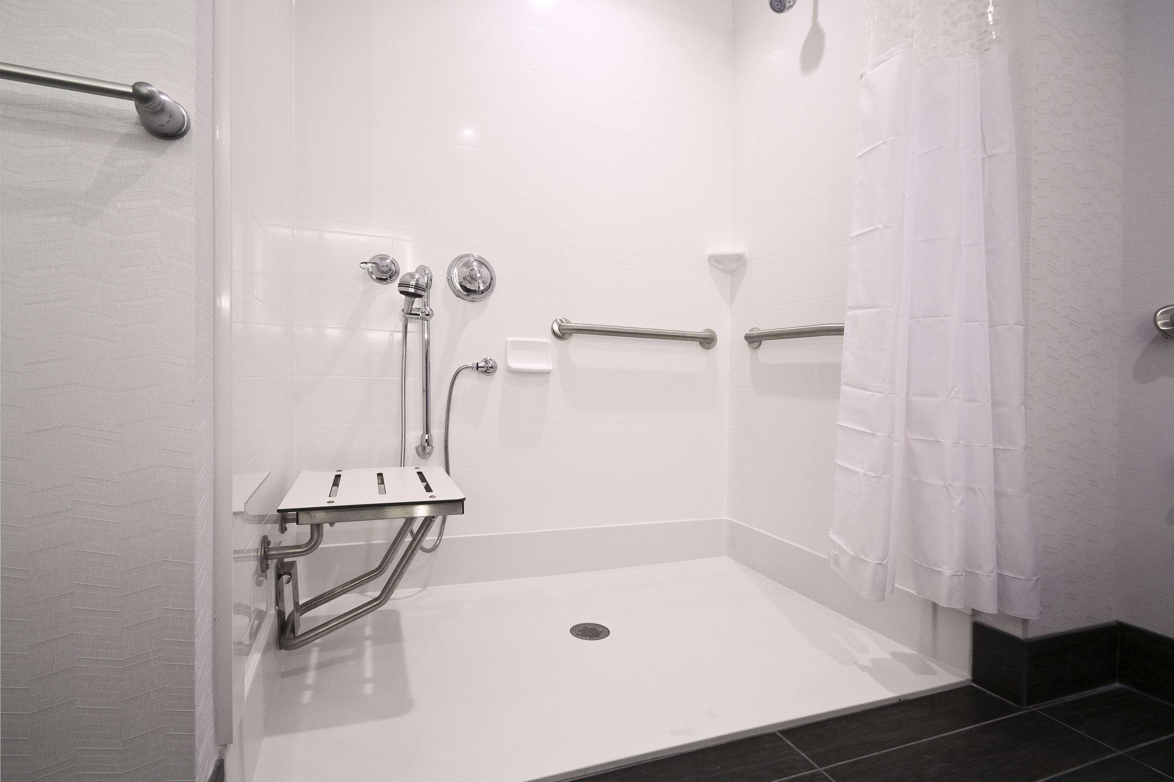 A modern shower area with a white tiled wall, a grab bar, a handheld showerhead, and a shower control panel.