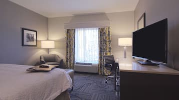 Ett hotellrum med en säng, två stolar, ett skrivbord med en tv och ett fönster med gardiner.