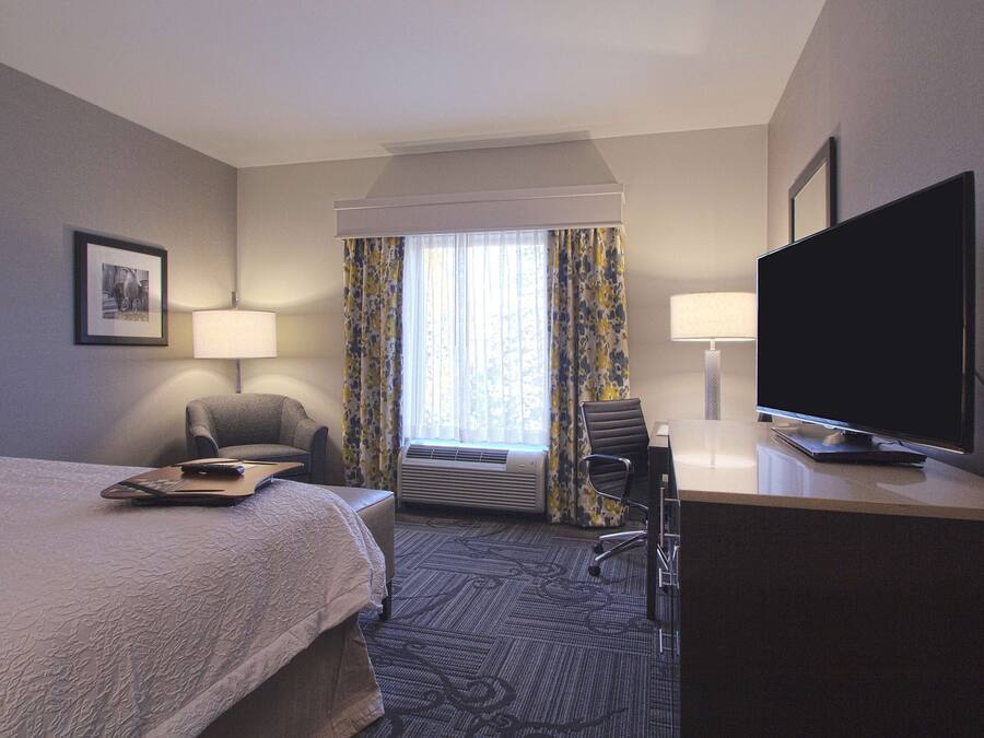 Ett hotellrum med en säng, två stolar, ett skrivbord med en tv och ett fönster med gardiner.
