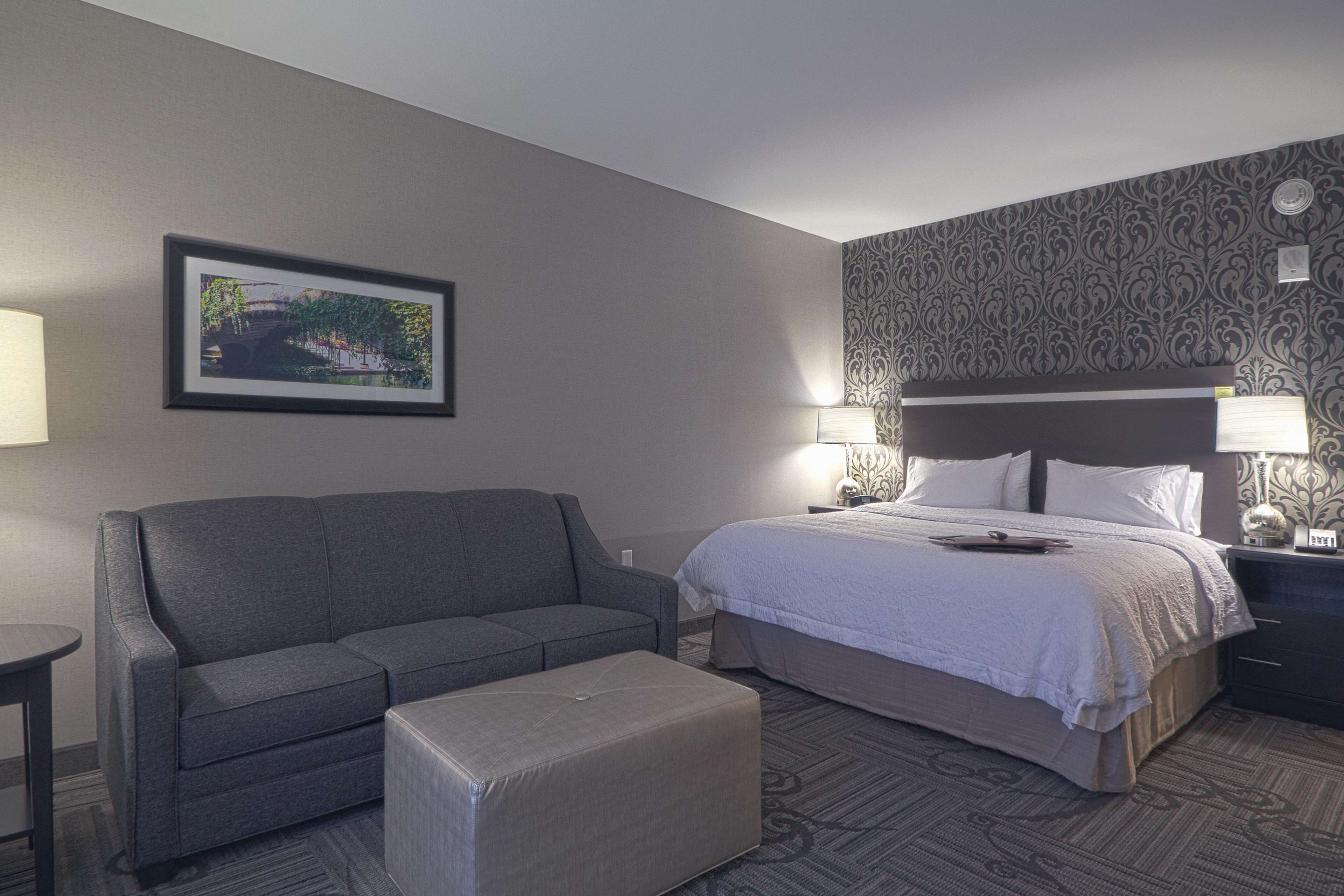 A hotel room with a bed, a sofa, an ottoman, a nightstand, and a wall-mounted lamp.