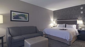 A hotel room with a bed, a sofa, an ottoman, a nightstand, and a wall-mounted lamp.