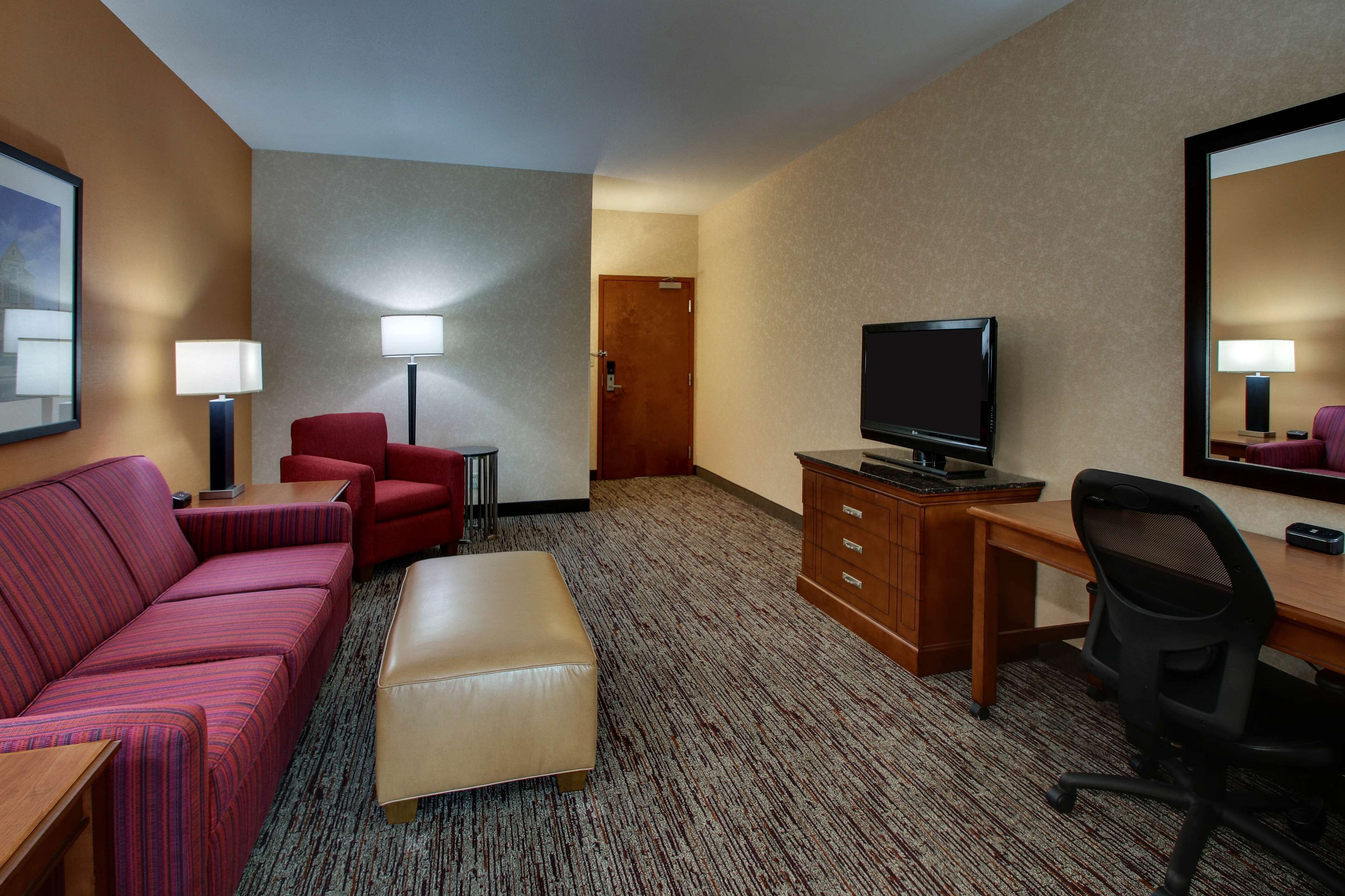 A hotel room with a sofa, ottoman, desk, chair, television, and a mirror.