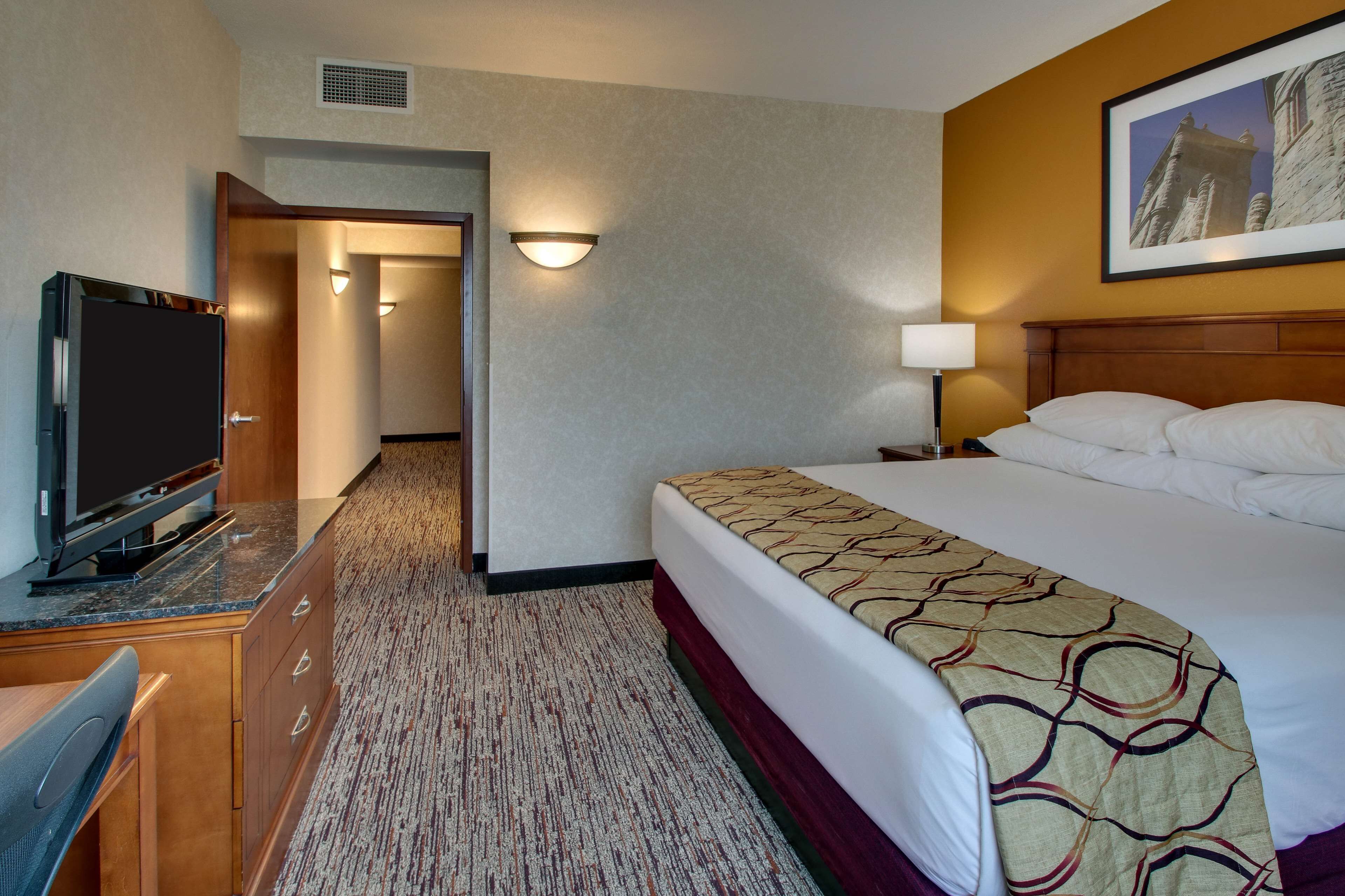 A hotel room with a bed, a desk with a television, a lamp, and a hallway leading to another room.