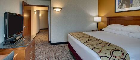 A hotel room with a bed, a desk with a television, a lamp, and a hallway leading to another room.