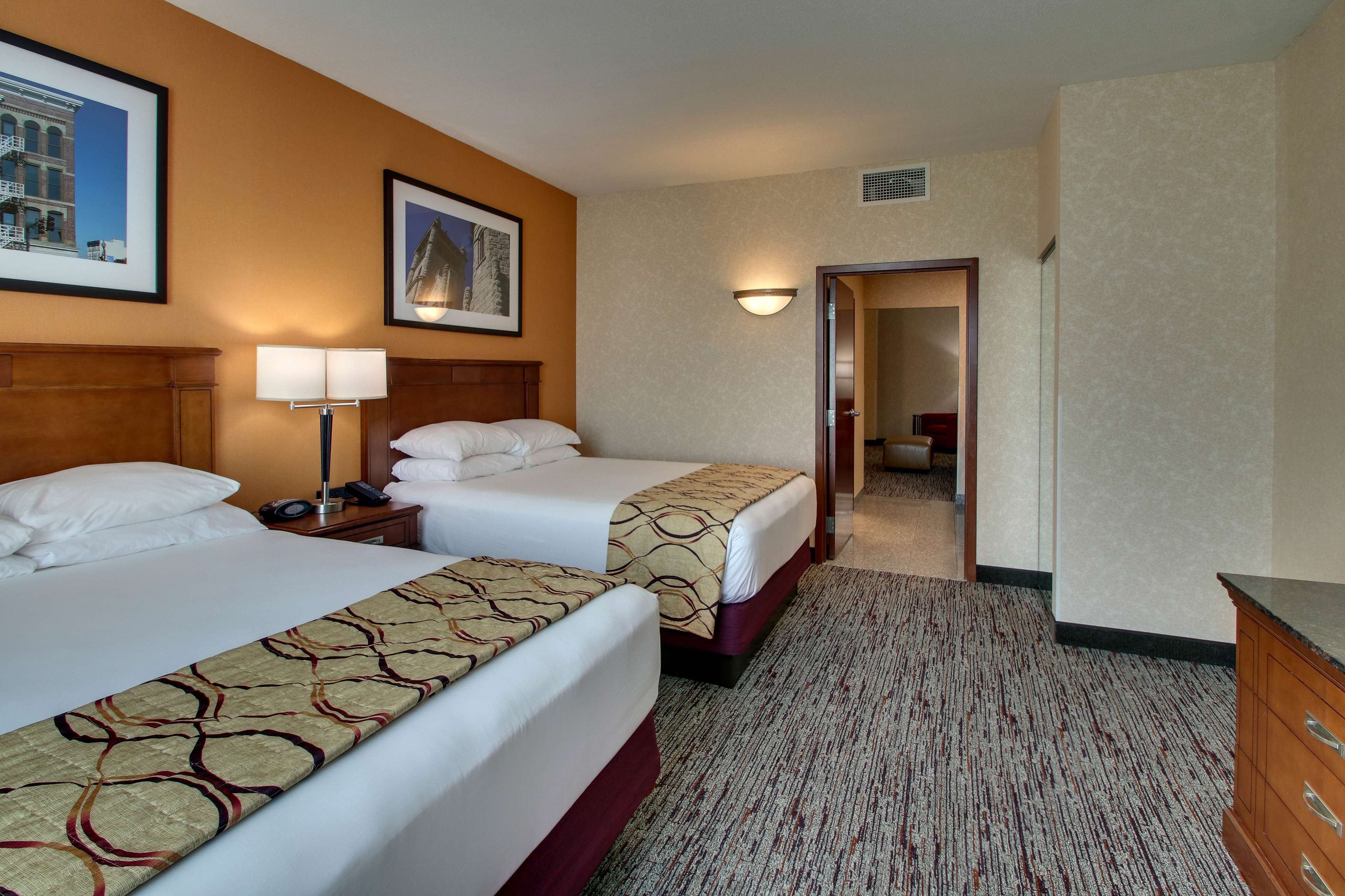 Ein Hotelzimmer mit zwei Betten, einem Nachttisch mit Lampe, einem Schminktisch und einem Flur, der zu einem anderen Zimmer führt.