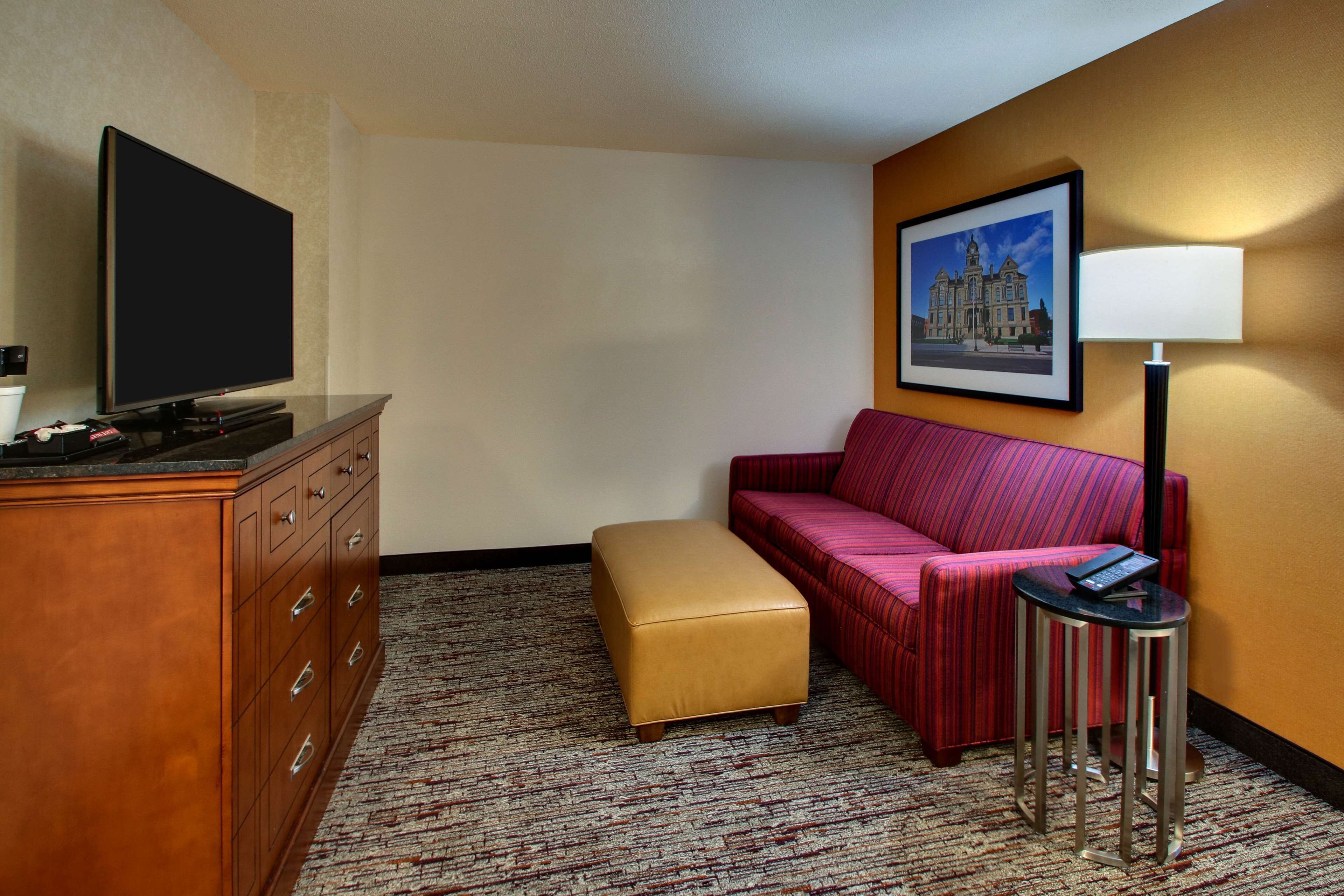 Una habitación de hotel con una televisión de pantalla plana sobre un aparador de madera, un sofá a rayas rojas, un pequeño reposapiés, una lámpara de pie y un cuadro enmarcado en la pared.