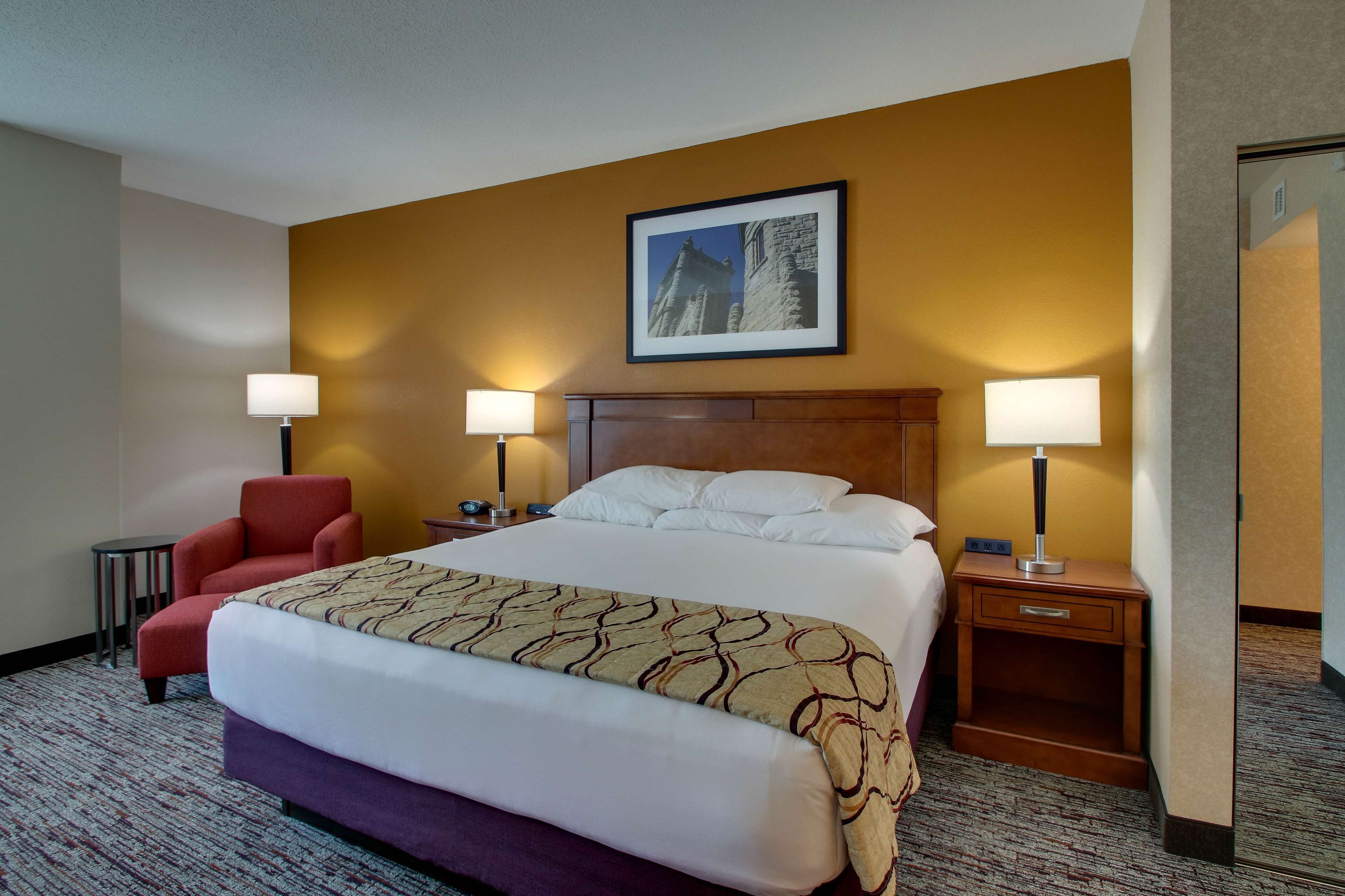 Une chambre d’hôtel avec un grand lit, deux tables de chevet avec des lampes, un fauteuil rouge et un tableau encadré au mur.