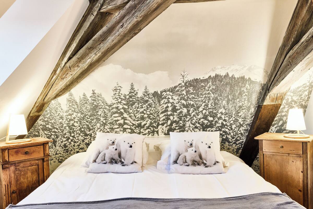 A bedroom with a high ceiling and exposed wooden beams, a bed with white bedding, two bedside tables with lamps, and a wall mural of a snowy forest.