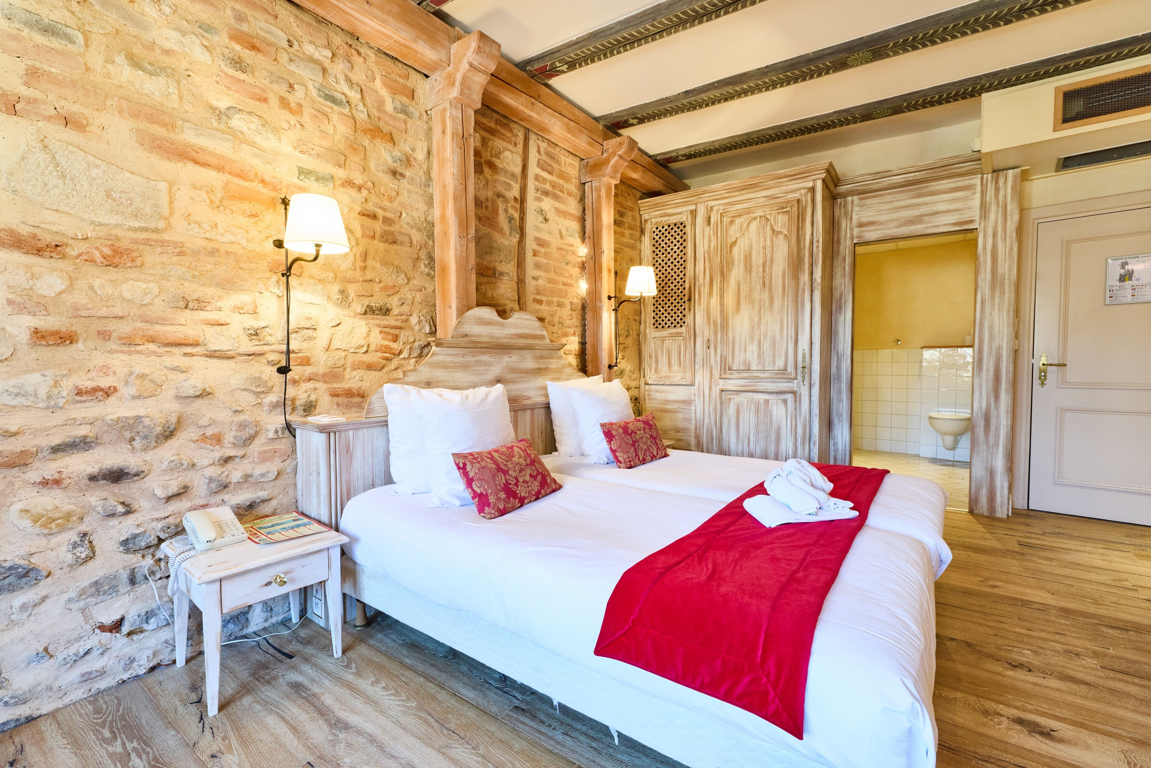 A large bed with white linens and a red throw blanket, a wooden nightstand with a lamp, and a stone wall.