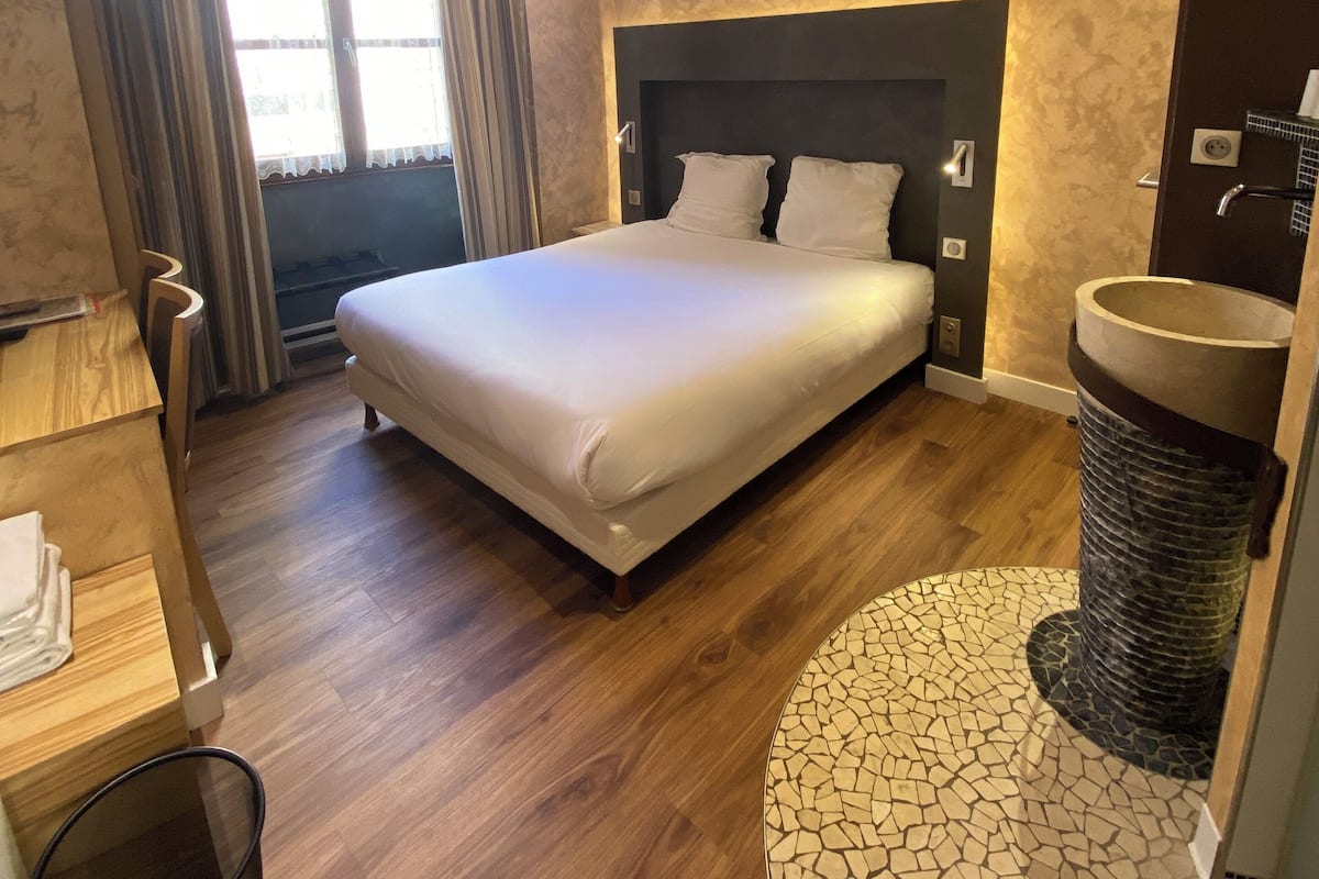 A bedroom with a large bed, wooden flooring, a desk, and a bathroom with a mosaic floor.
