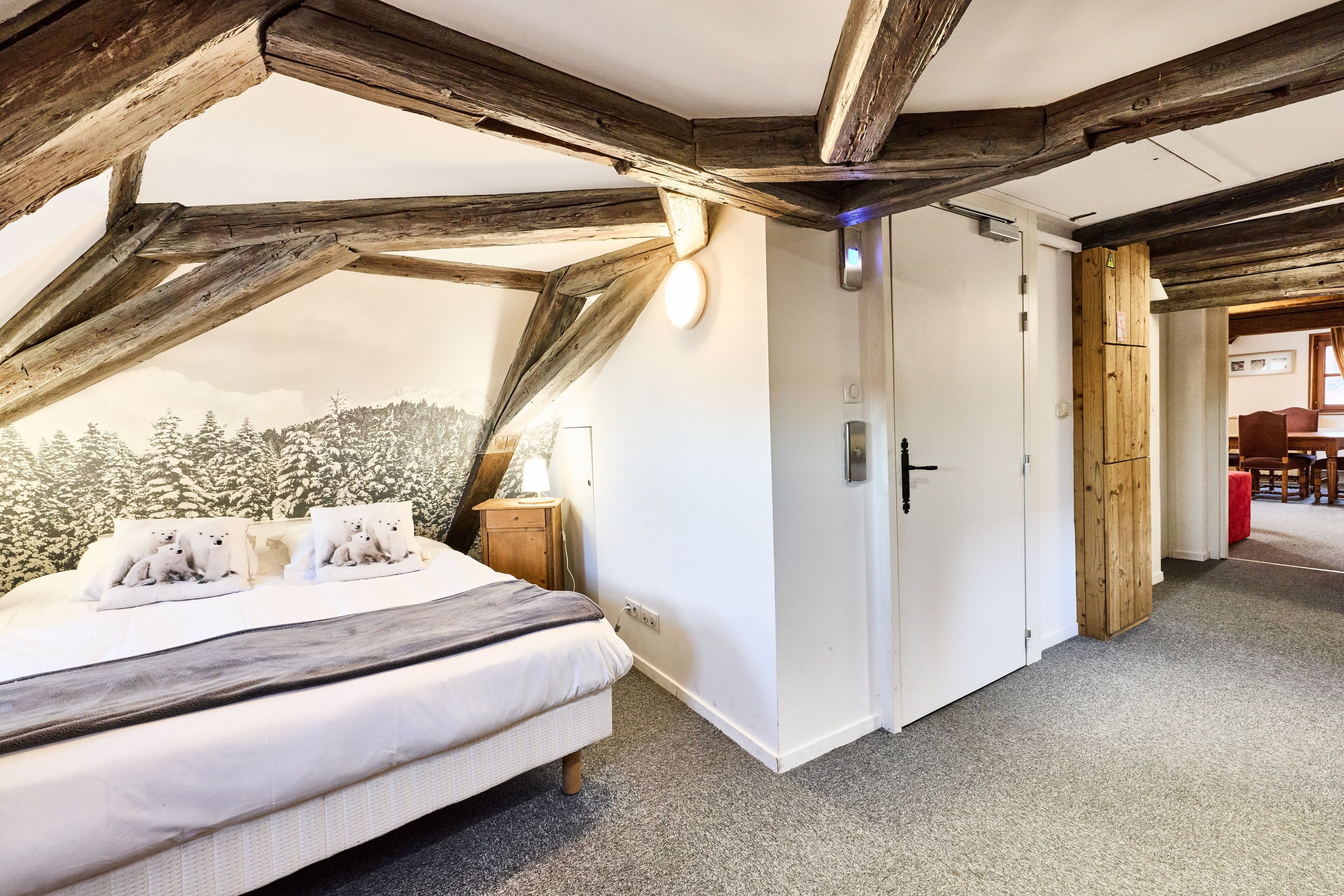 A bedroom with a bed, wooden beams, a wardrobe, and a view of trees.