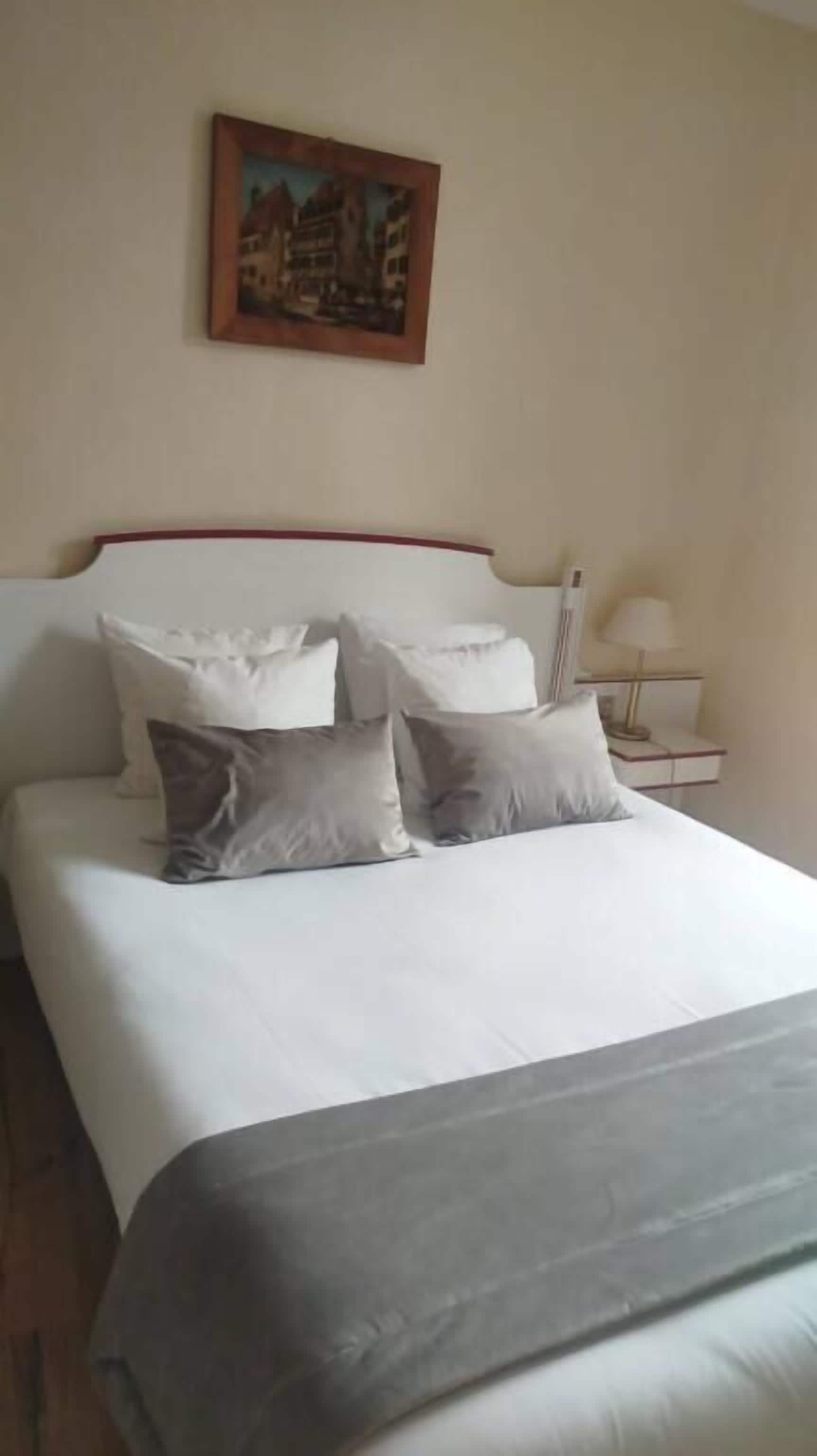 A neatly made bed with white and grey bedding, a red headboard, and a painting of buildings on the wall.
