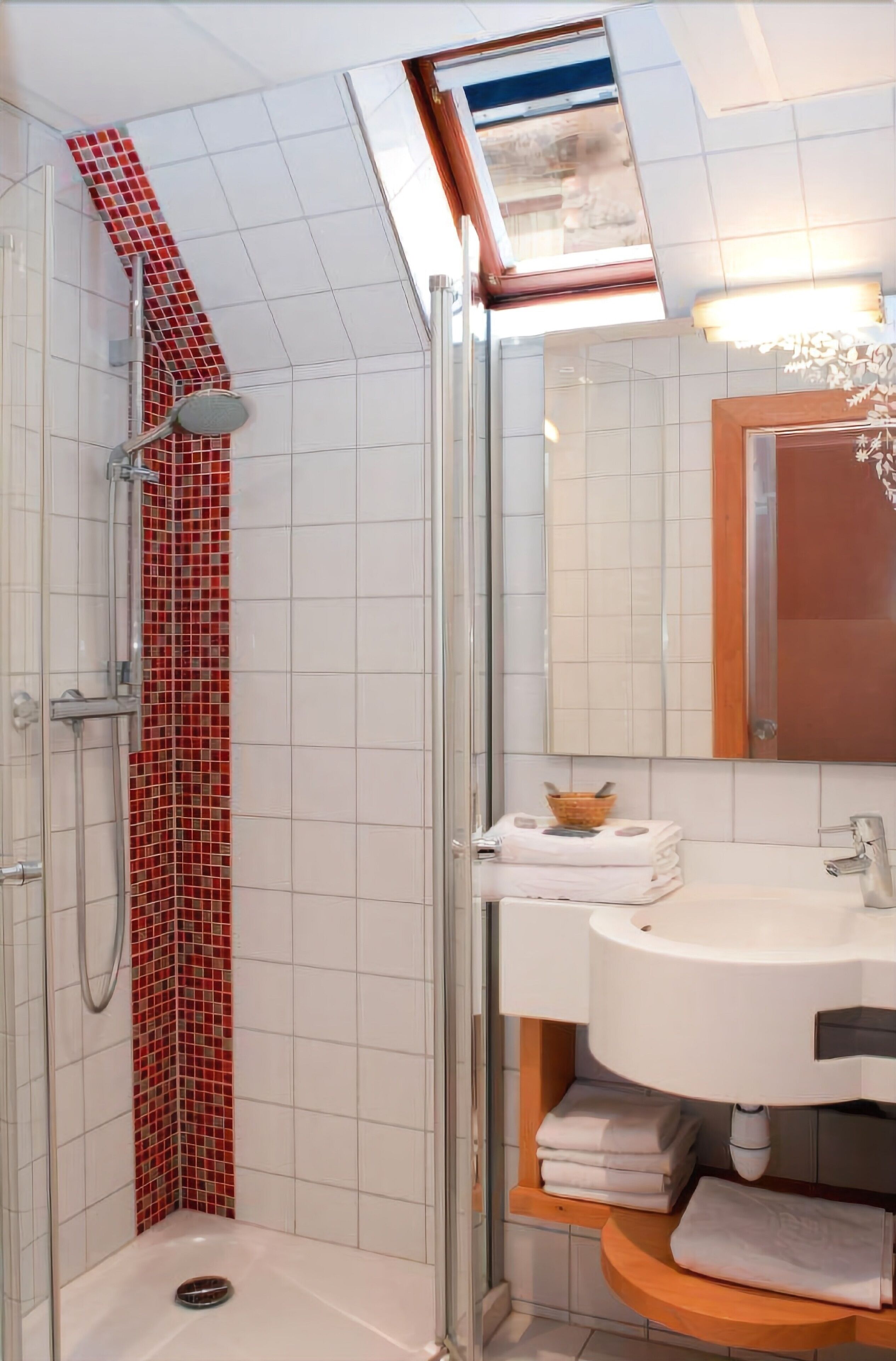 Un baño moderno con ducha acristalada, un lavamanos blanco sobre un mueble de madera y pared de ducha con azulejos rojos.