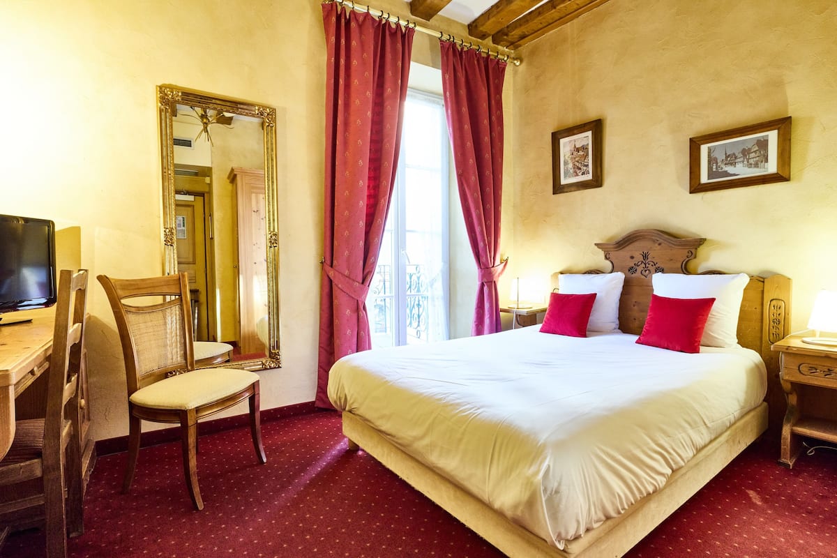 A hotel room with a large bed, a wooden chair, a desk with a television, and a large mirror.