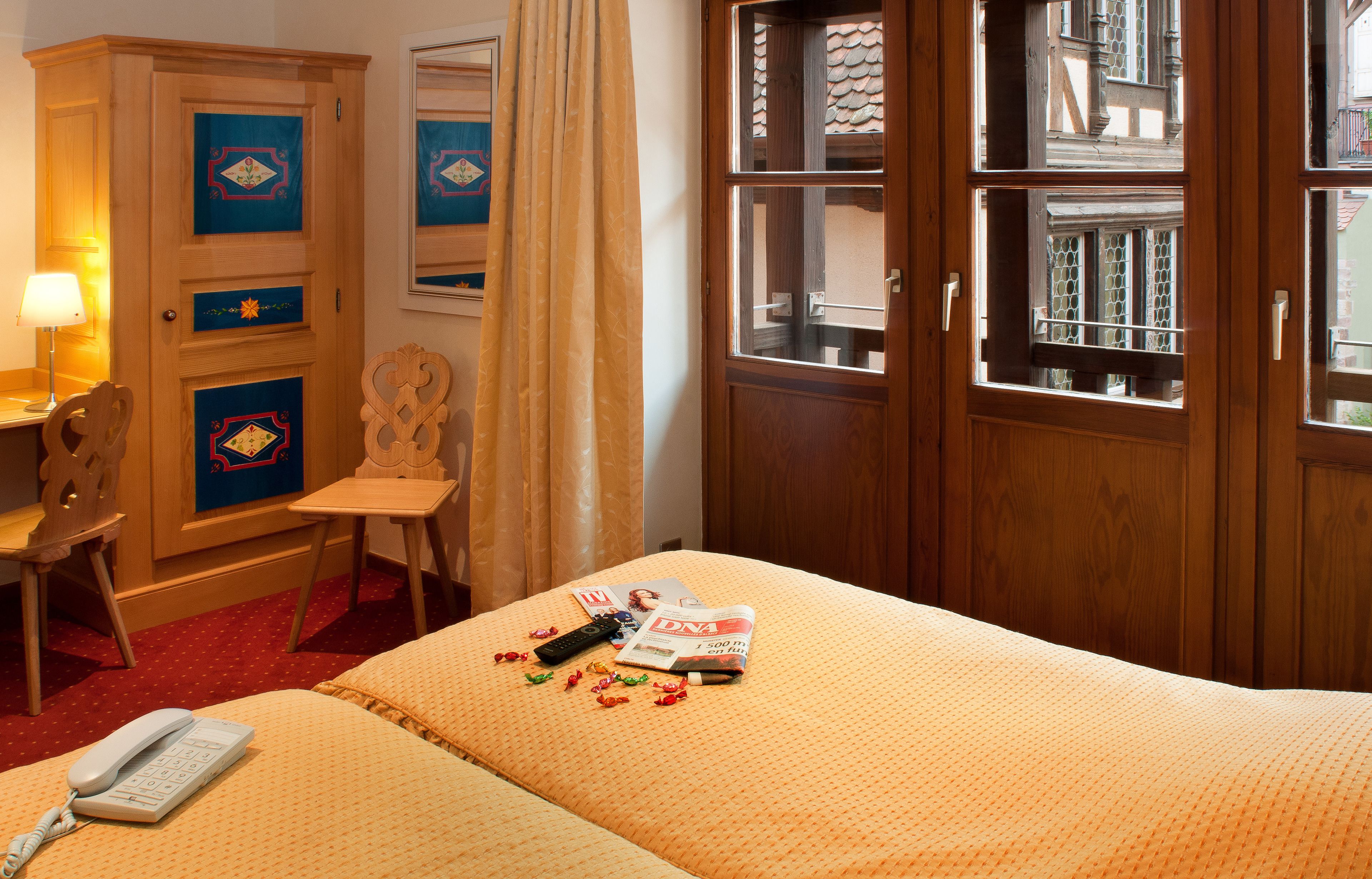 A hotel room with a bed, a wooden wardrobe, a desk with a lamp, a chair, and a window with a view of buildings.