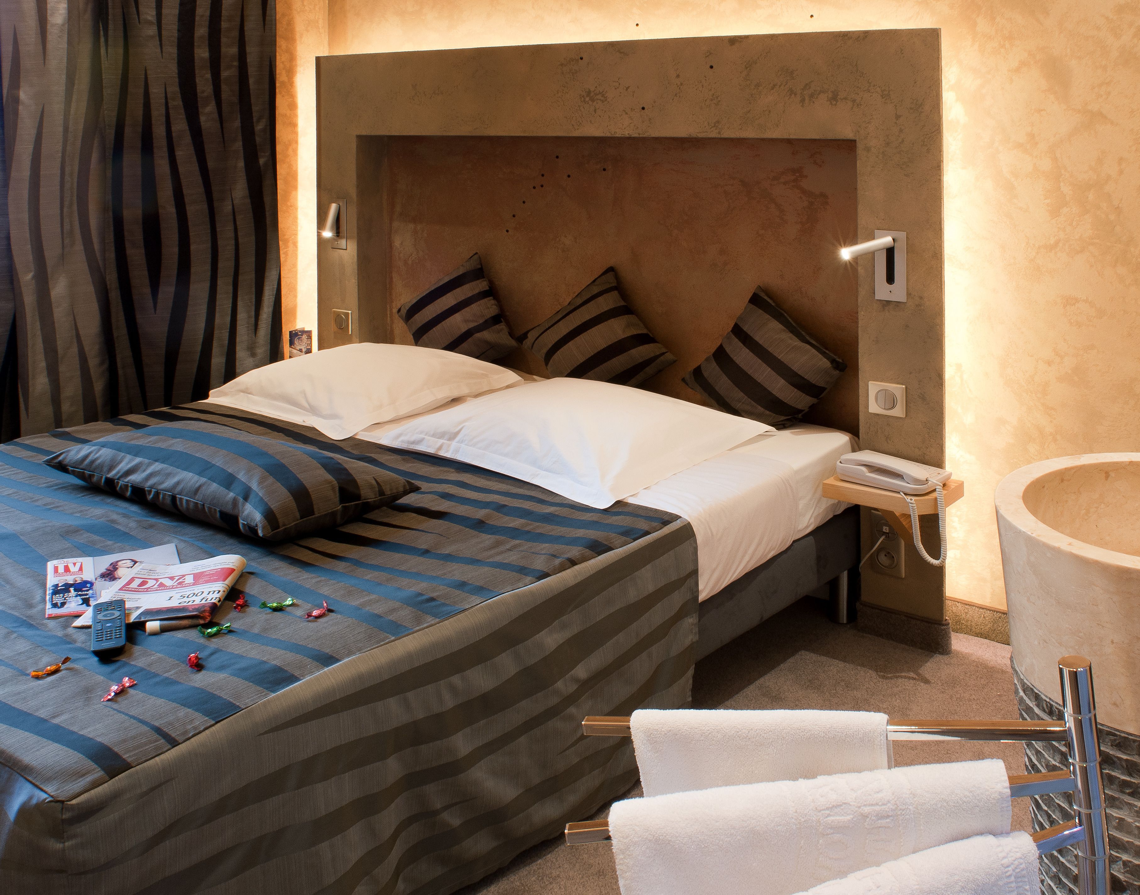 Quarto de hotel com cama, almofadas, mesa de cabeceira com telefone, revista e porta-toalhas.